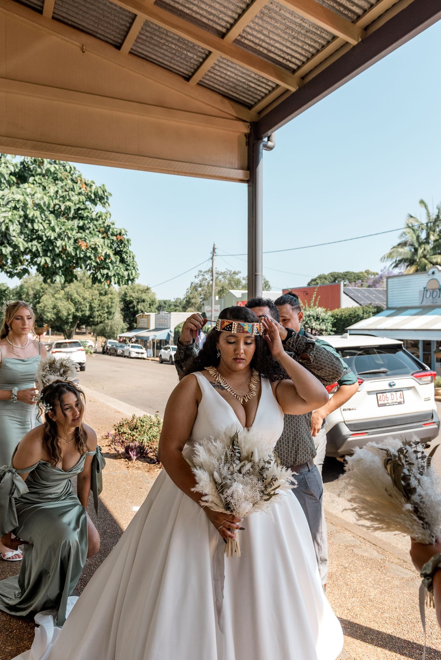 Bride in white gown with cultural headpiece holds bouquet while bridesmaids in sage green assist before wedding ceremony.