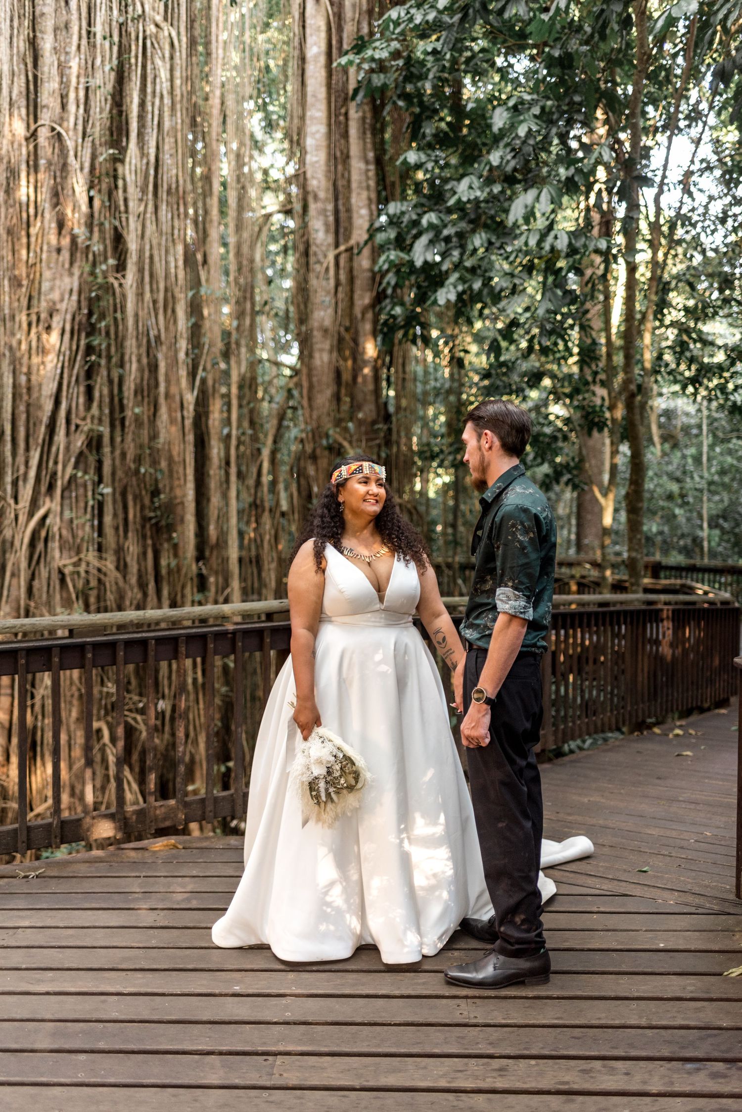 Bride and groom holding hands and smiling on a wooden deck with banyan tree forest backdrop.