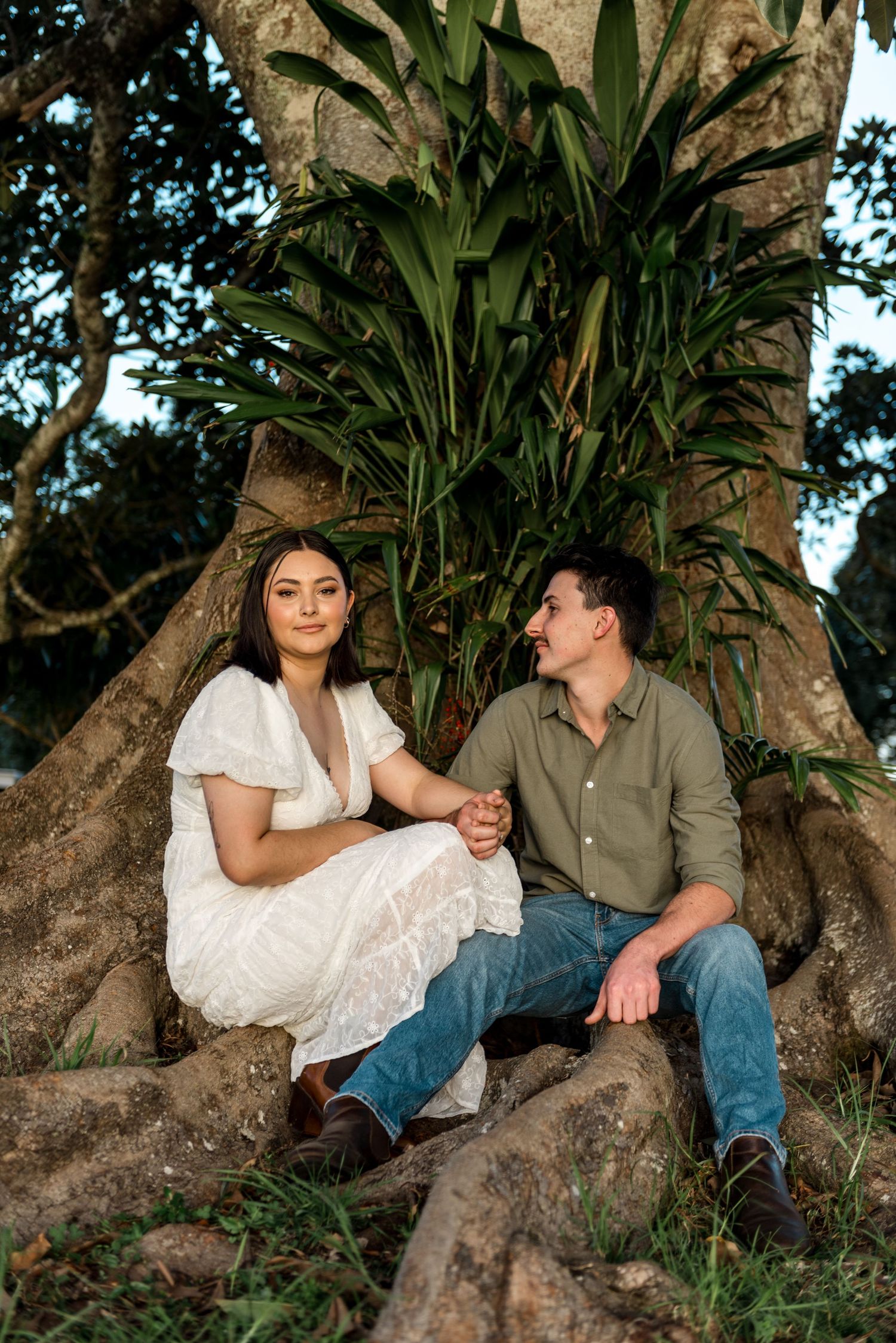 Couple sitting at base of large tree holding hands, woman in white dress and man in green shirt.