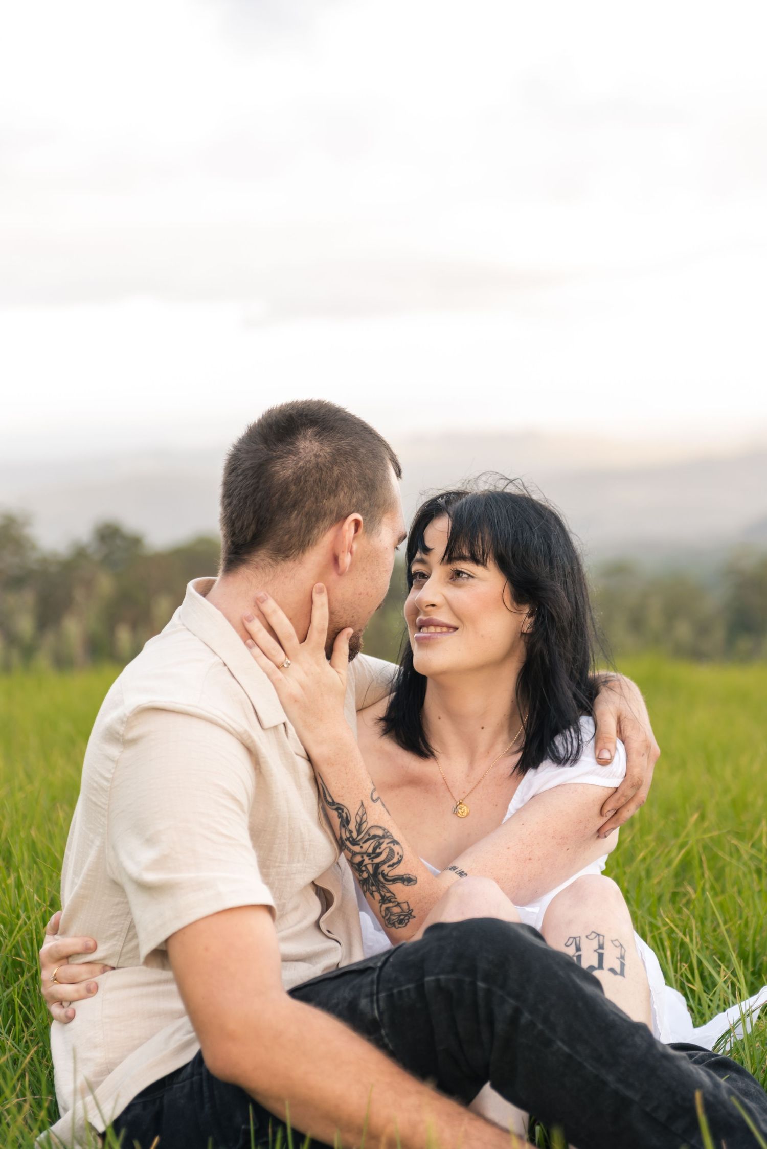 Couple sitting in green field, woman smiling and touching mans face, both embracing romantically outdoors.