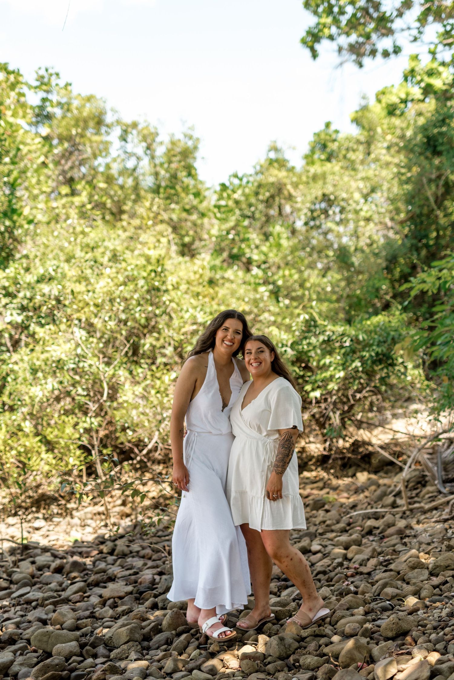 Two smiling women in white dresses stand together on a rocky path surrounded by vibrant green tropical foliage.