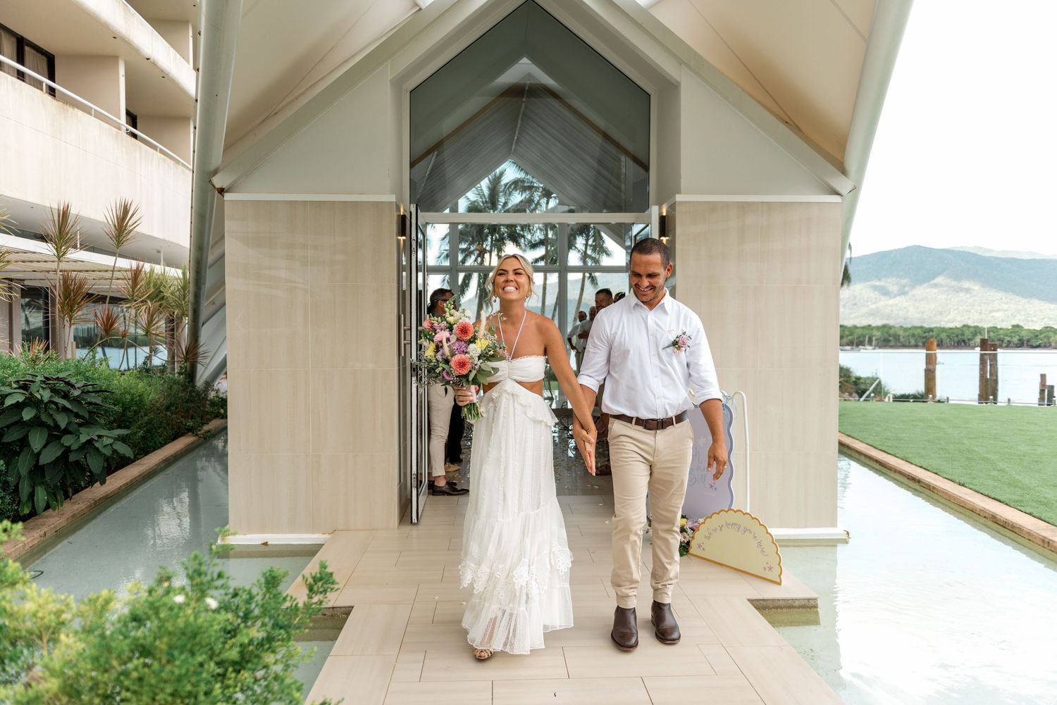 Bride and groom walk hand in hand from a chapel at a tropical waterfront wedding venue.