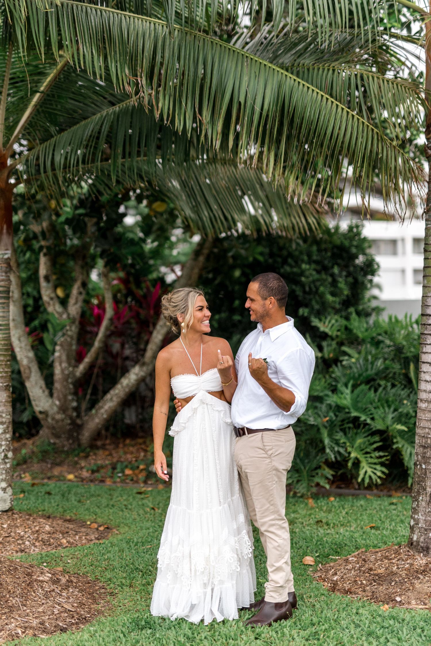 Bride and groom laughing together on green grass beneath a palm tree at a tropical wedding location.