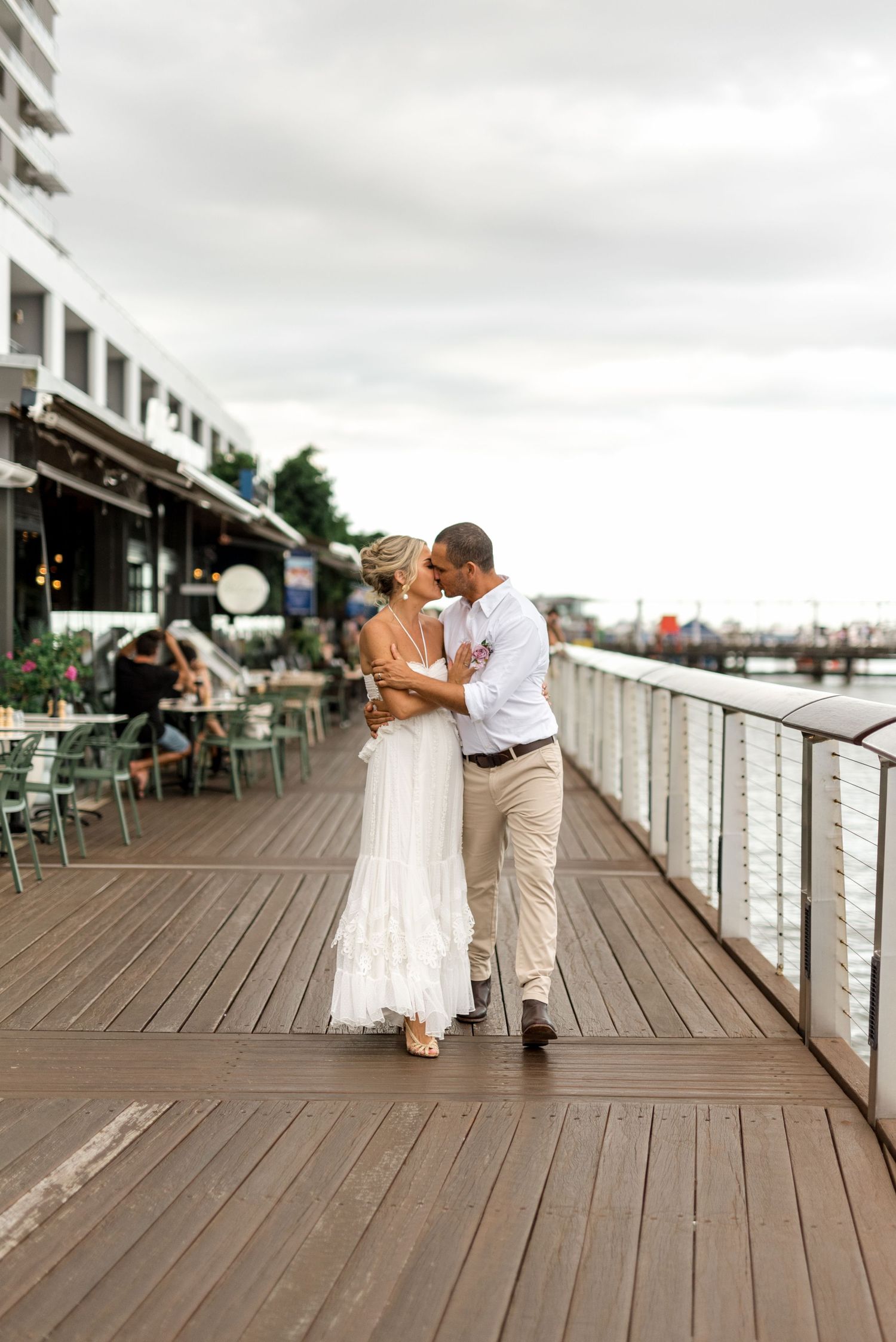 Bride and groom share a romantic kiss while strolling along a waterfront boardwalk at dusk.