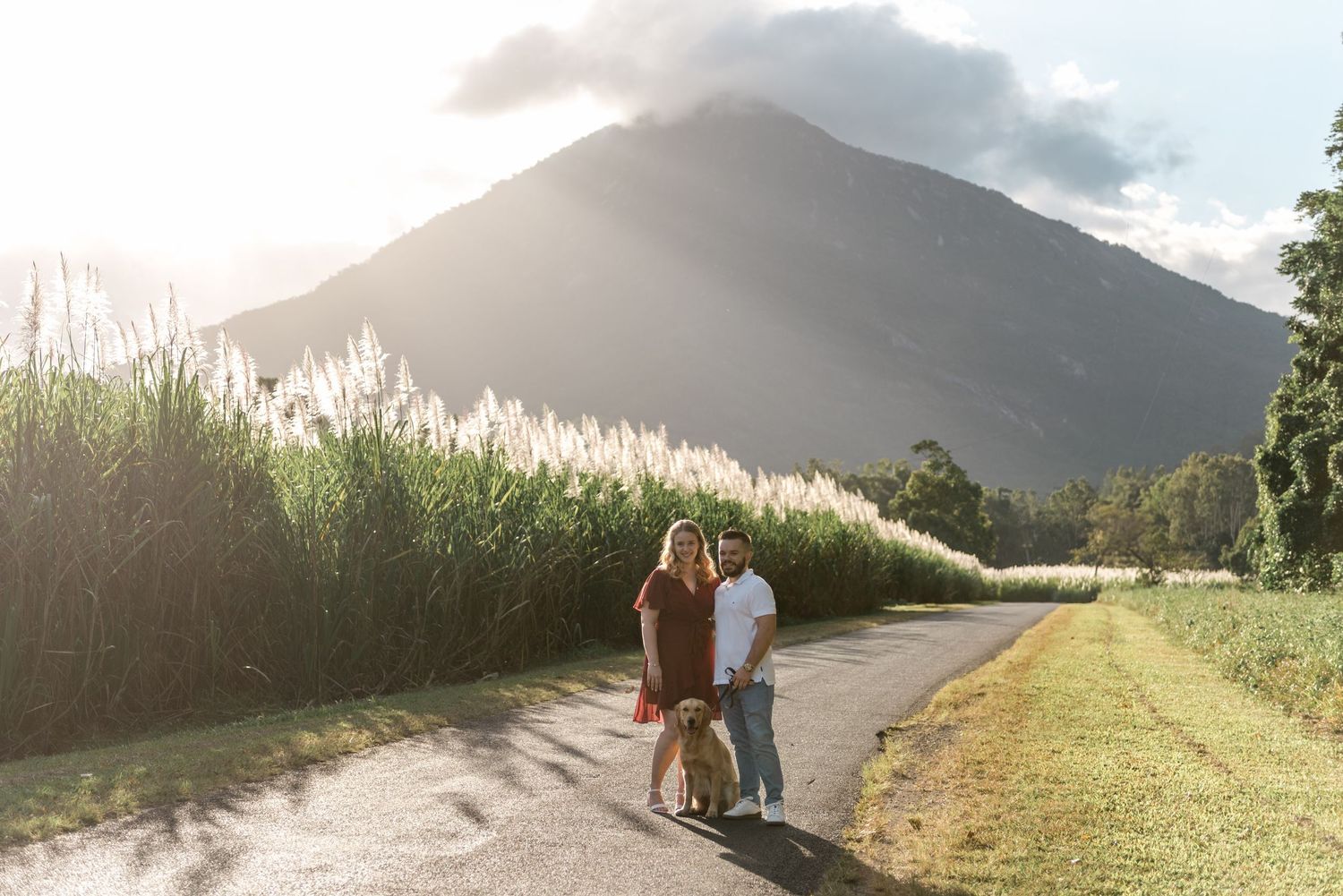 Couple with golden retriever standing on scenic rural road with mountain backdrop and golden hour sunlight.