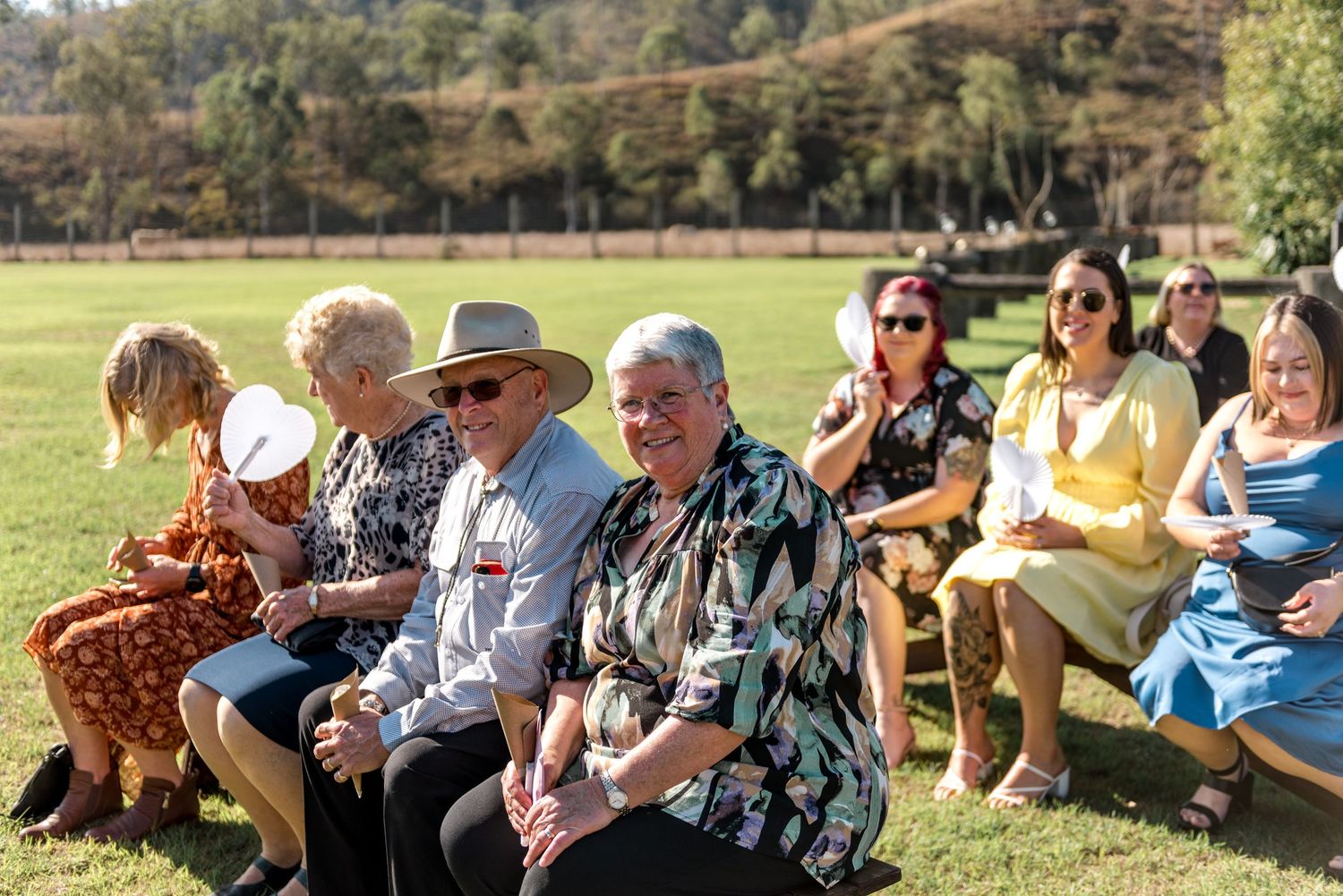 Wedding guests seated outdoors on sunny day at rustic countryside wedding ceremony.