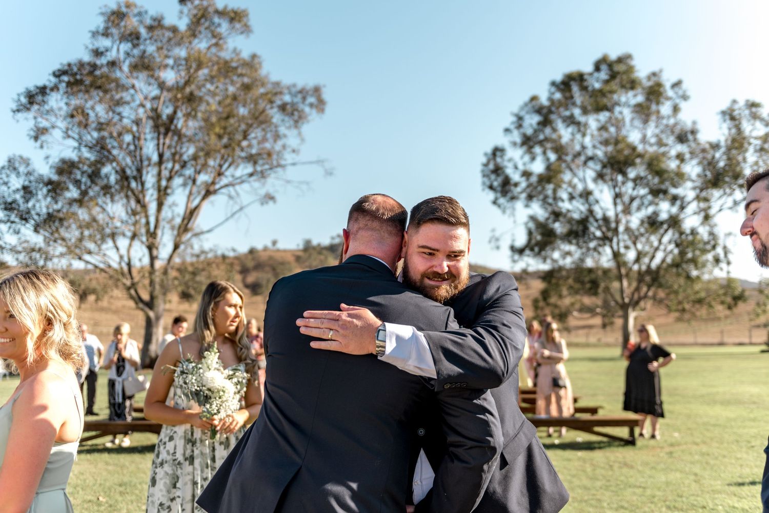 Two groomsmen share a warm embrace after outdoor wedding ceremony with guests mingling in background.