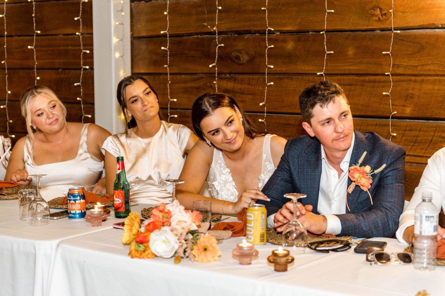 Bride and groom with bridesmaids seated at wedding head table with floral arrangements.
