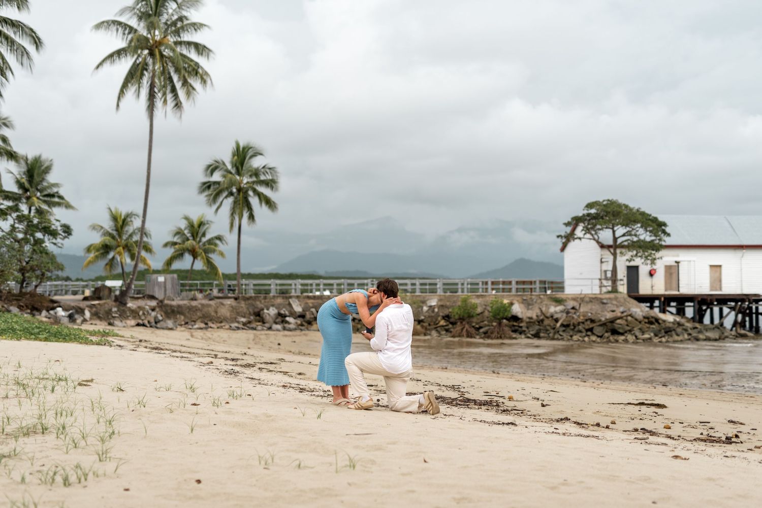 Man kneeling on tropical beach proposing to woman in blue dress near pier on cloudy day.