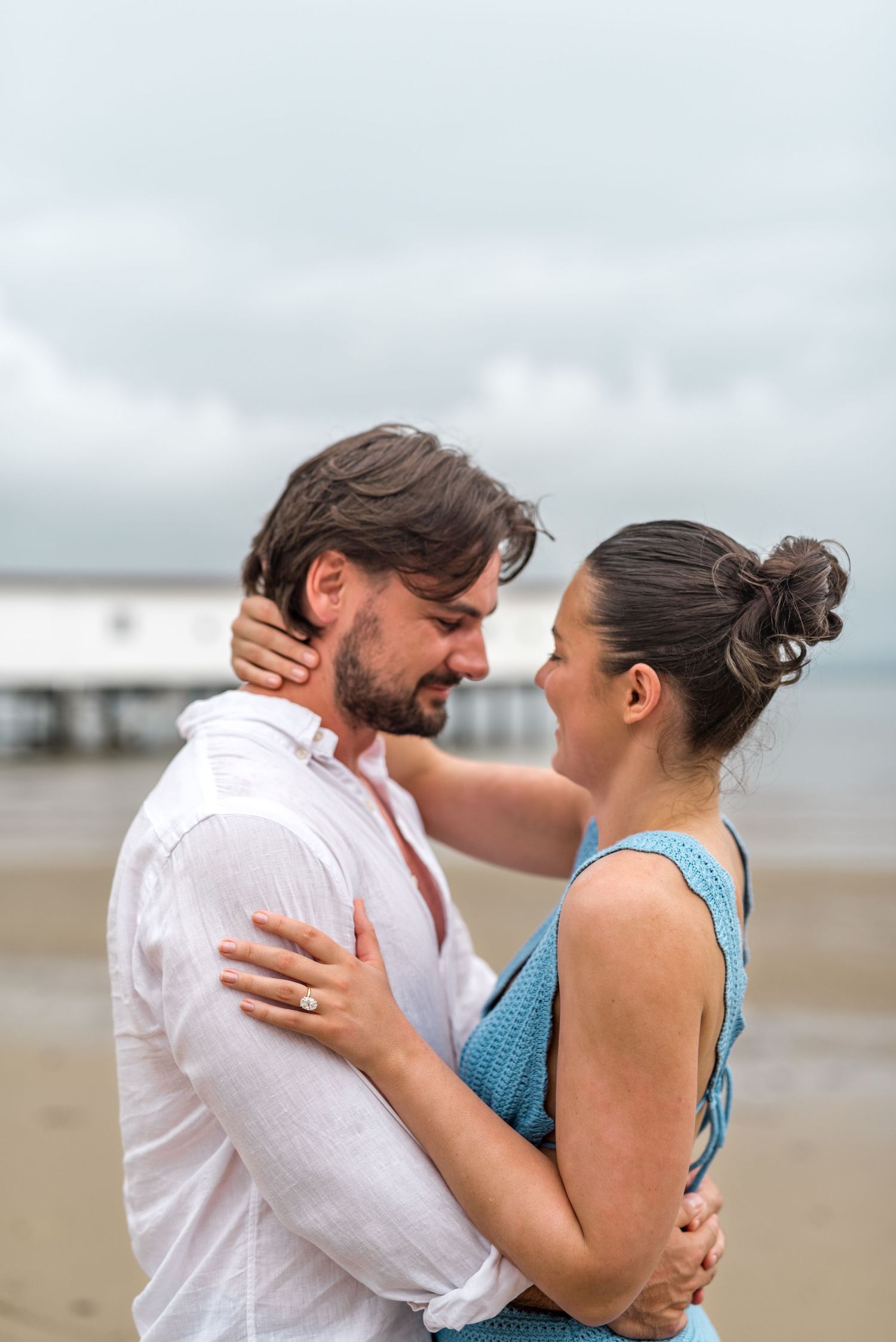 Couple embracing and smiling at each other on a beach with a pier visible behind them.