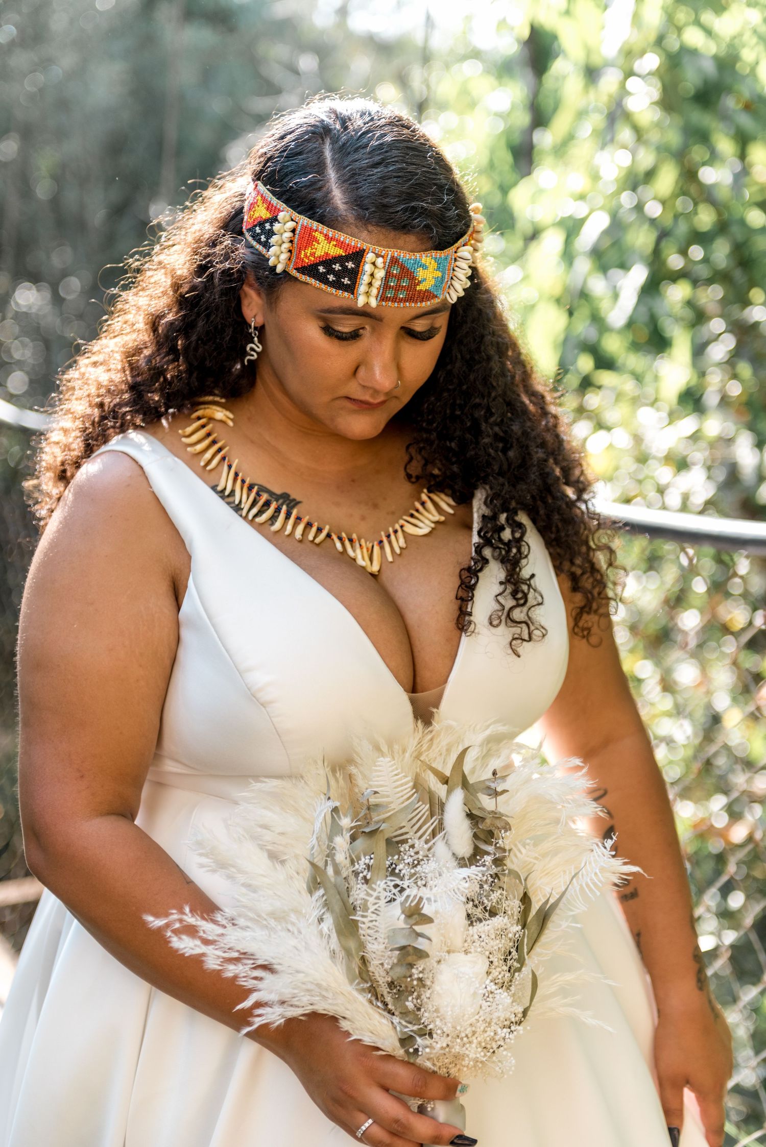 A bride in a white gown wears a colorful beaded headpiece and gold necklace outdoors.