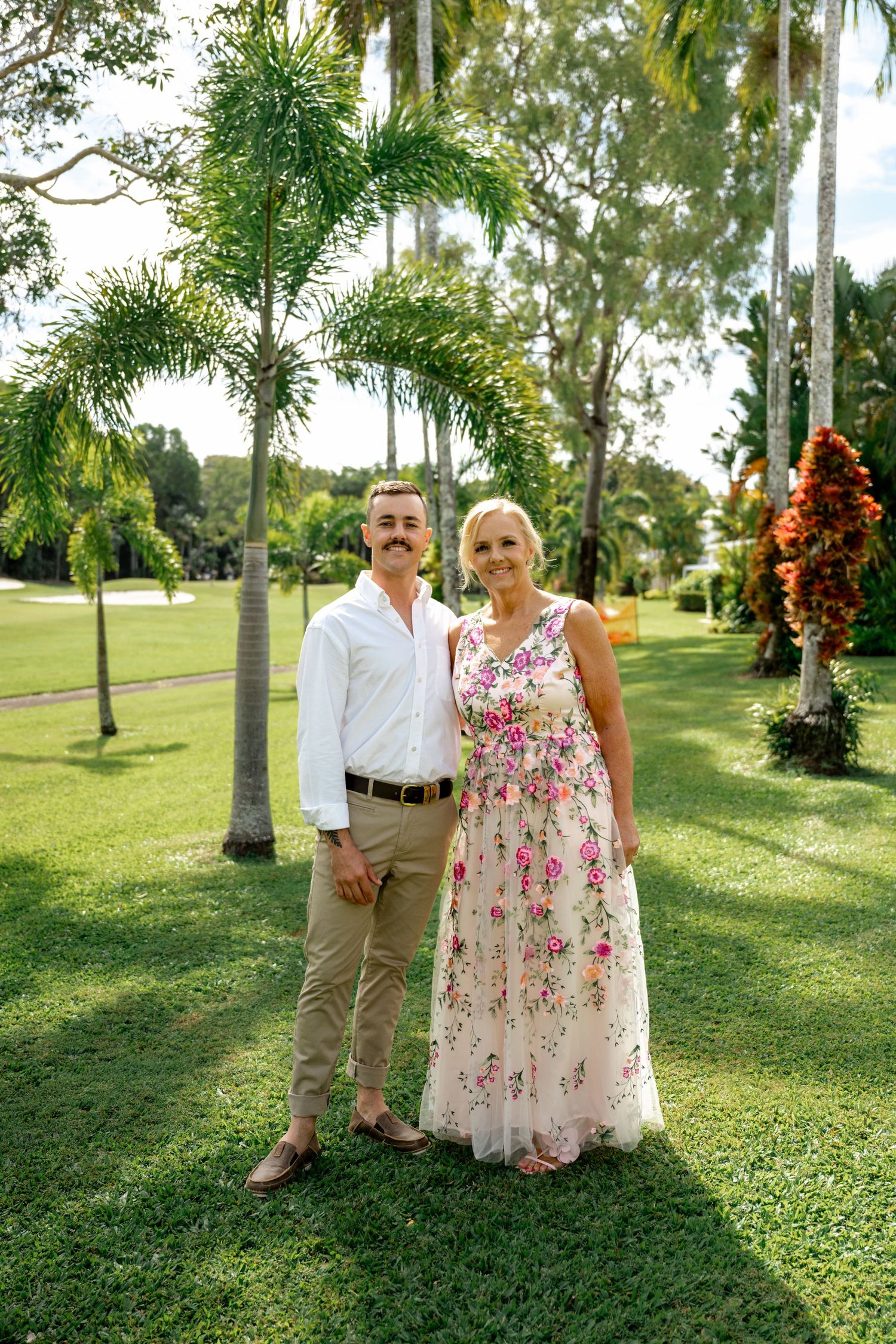 Man in white shirt and woman in floral maxi dress posing together in a tropical garden.