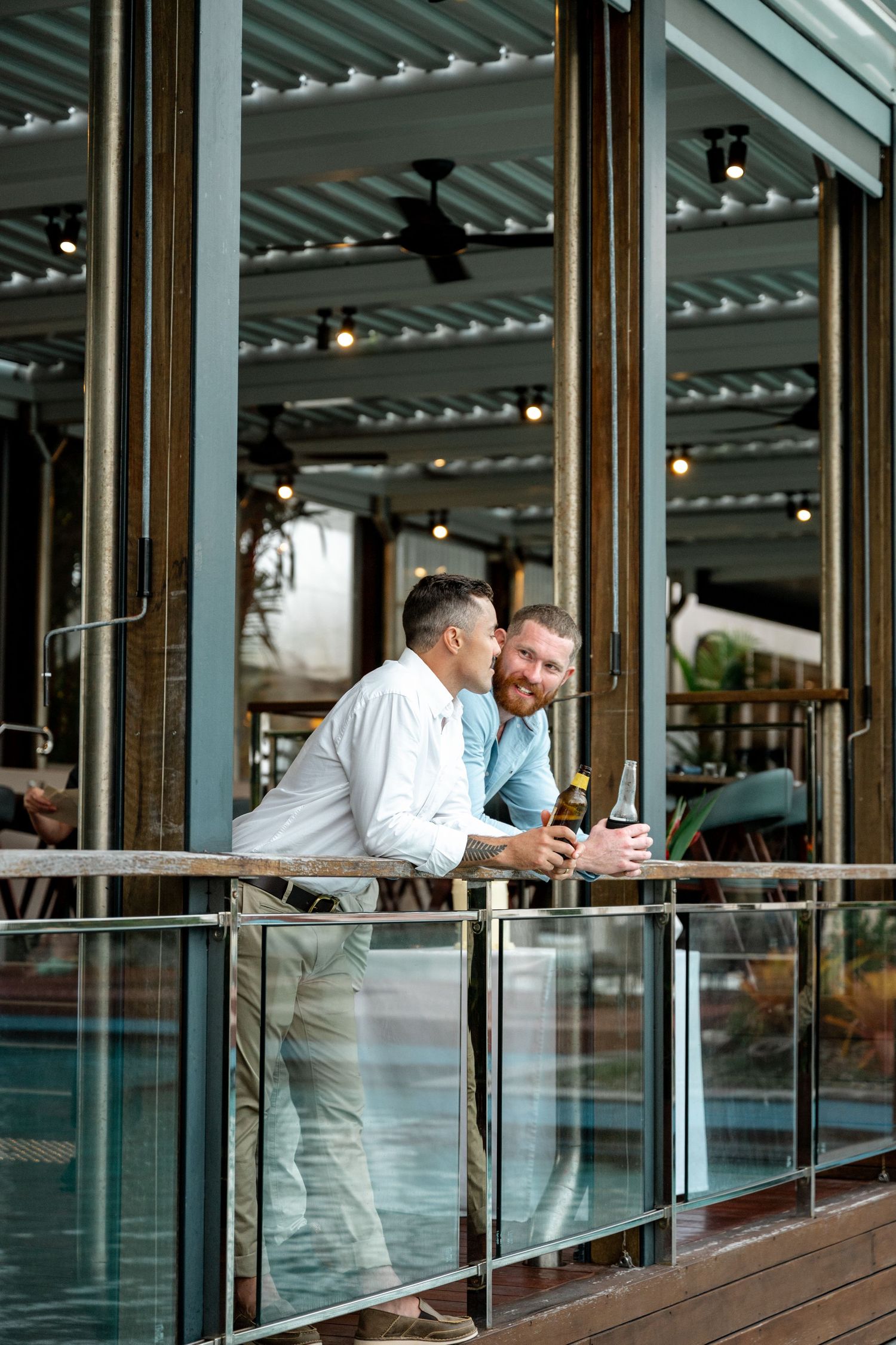 Two men in smart casual attire enjoying drinks on a balcony overlooking a tropical resort pool at Sheraton Grand Mirage Port Douglas.