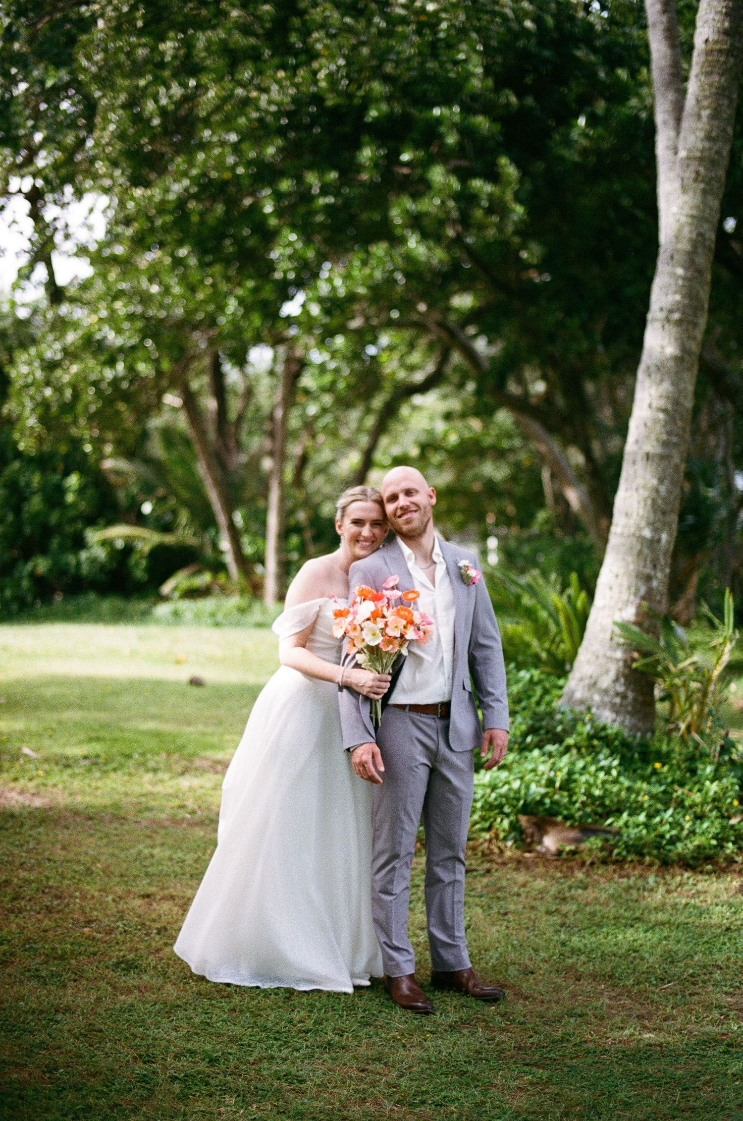 Smiling bride and groom pose together on green lawn with tropical trees in background at outdoor wedding.
