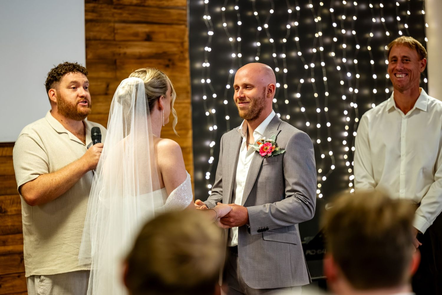 Groom in grey suit smiles at bride during wedding ceremony with fairy light backdrop and officiant holding microphone.