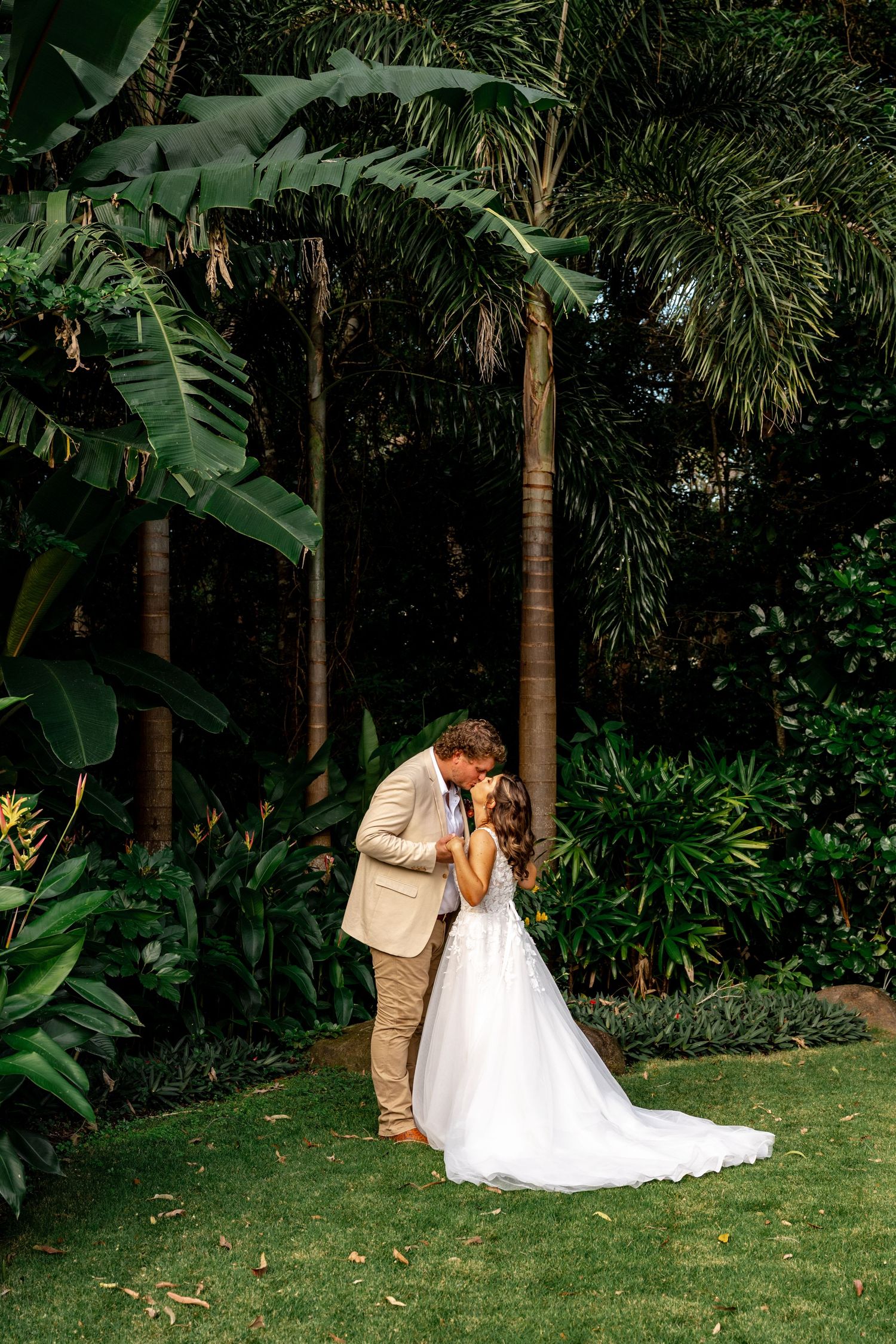 Bride and groom sharing first kiss surrounded by lush tropical palm trees in garden wedding setting.