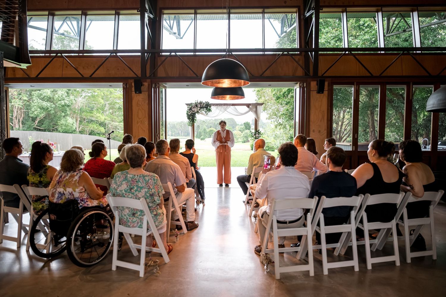 Wedding guests seated in white chairs watch a ceremony officiant in an open barn venue.