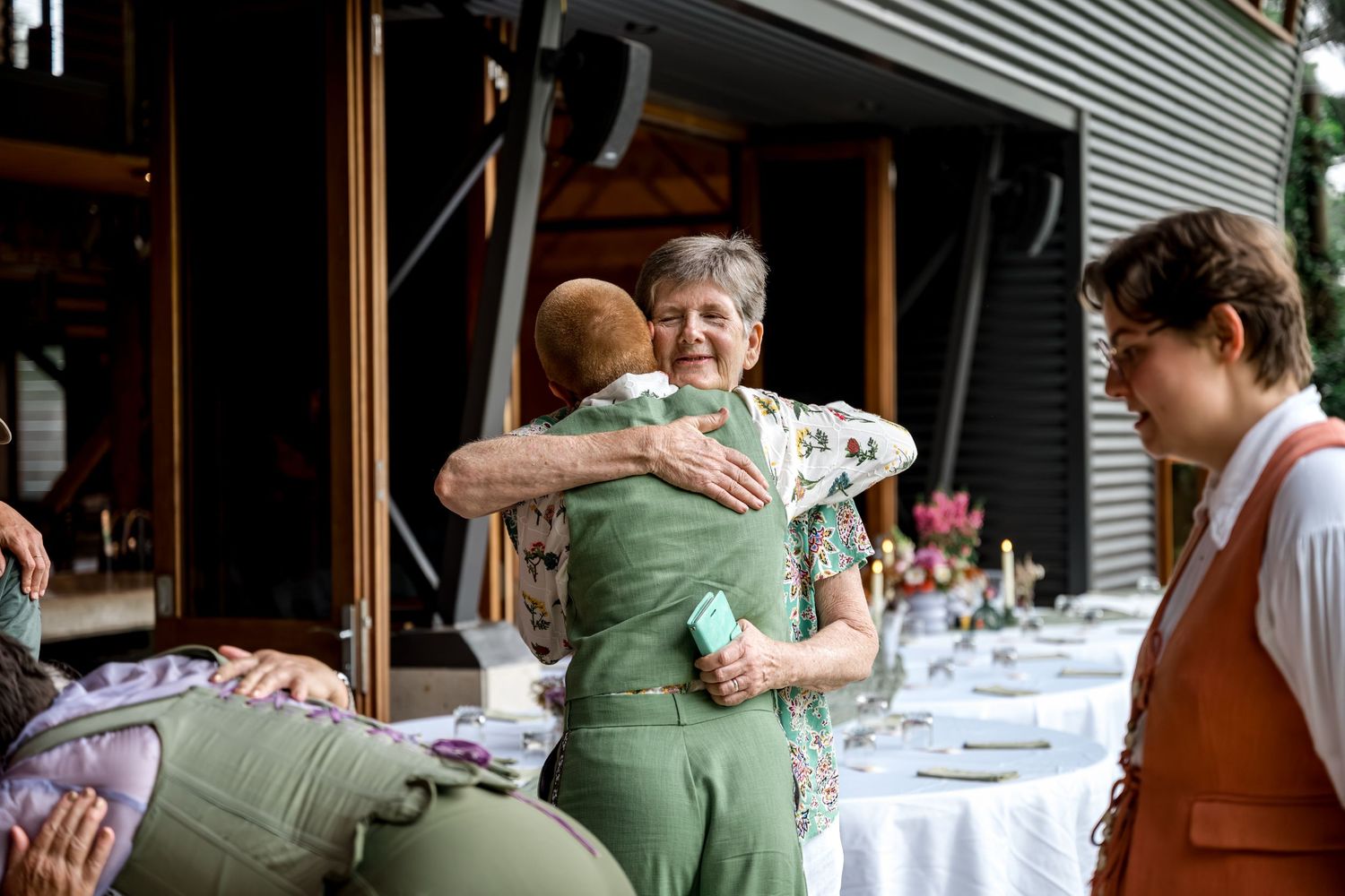 Elderly woman hugs a guest in a green suit at an outdoor wedding reception.