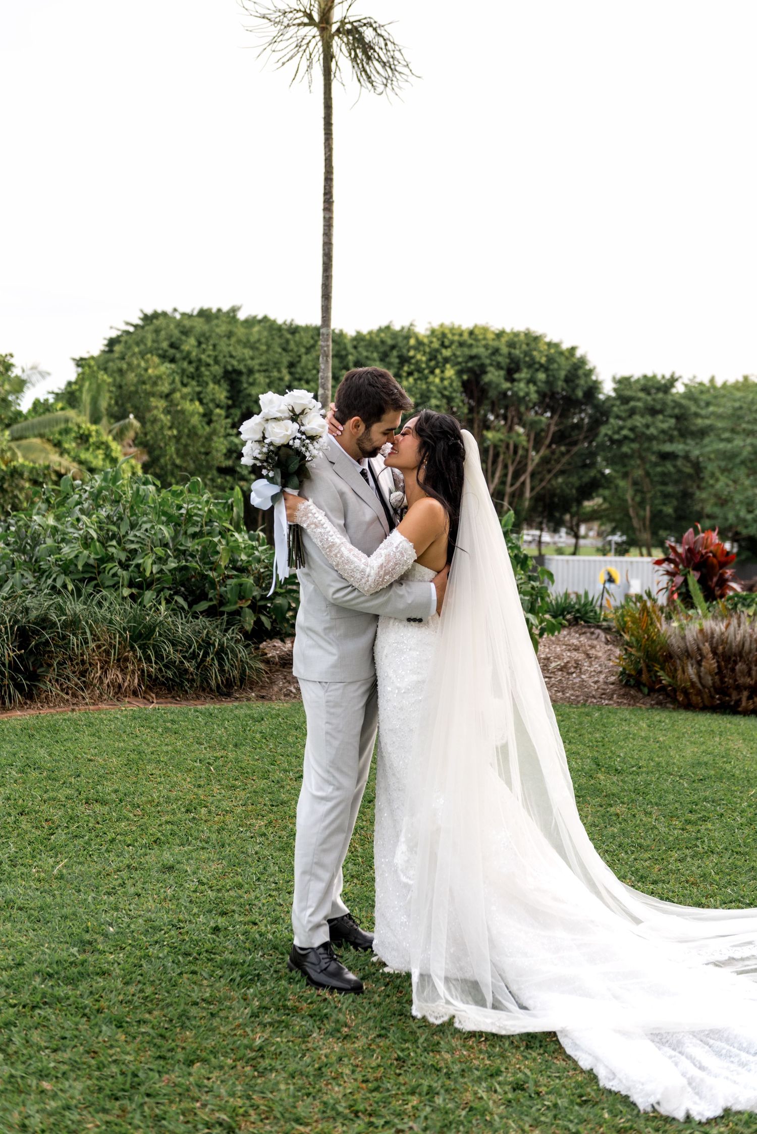 Bride and groom kissing in tropical garden, bride wearing long veil and white bouquet.