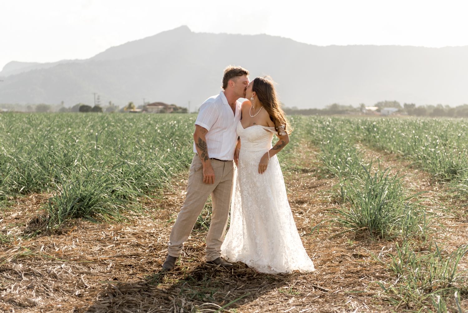 Groom tenderly kissing bride in white off-shoulder lace dress in a rustic outdoor field setting.