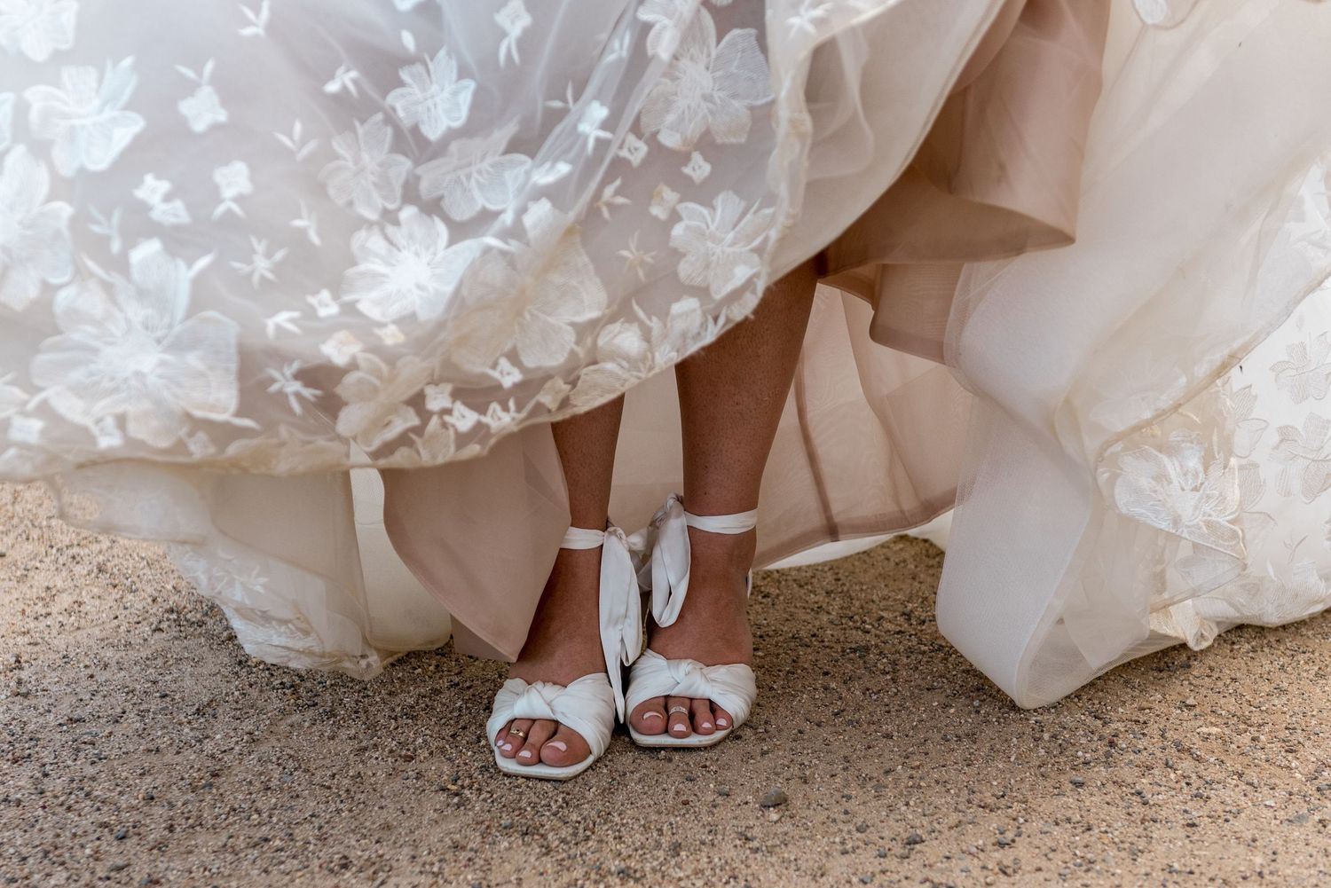 Close-up of bride wearing elegant white strappy heels beneath a lace and tulle wedding gown.