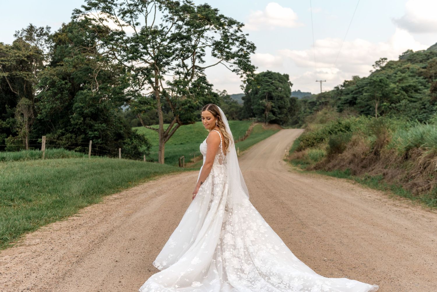 Bride in a floral lace wedding gown with long veil poses gracefully on a winding rural gravel road.