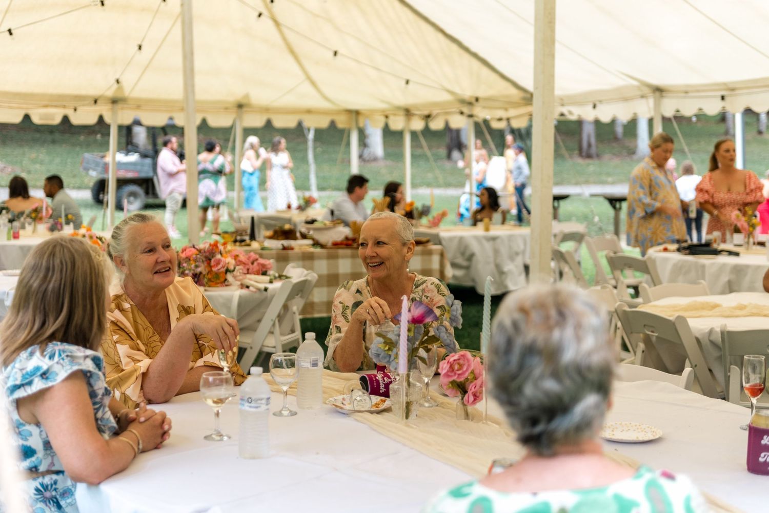 Wedding guests laughing and socializing at decorated tables under elegant white event tent.