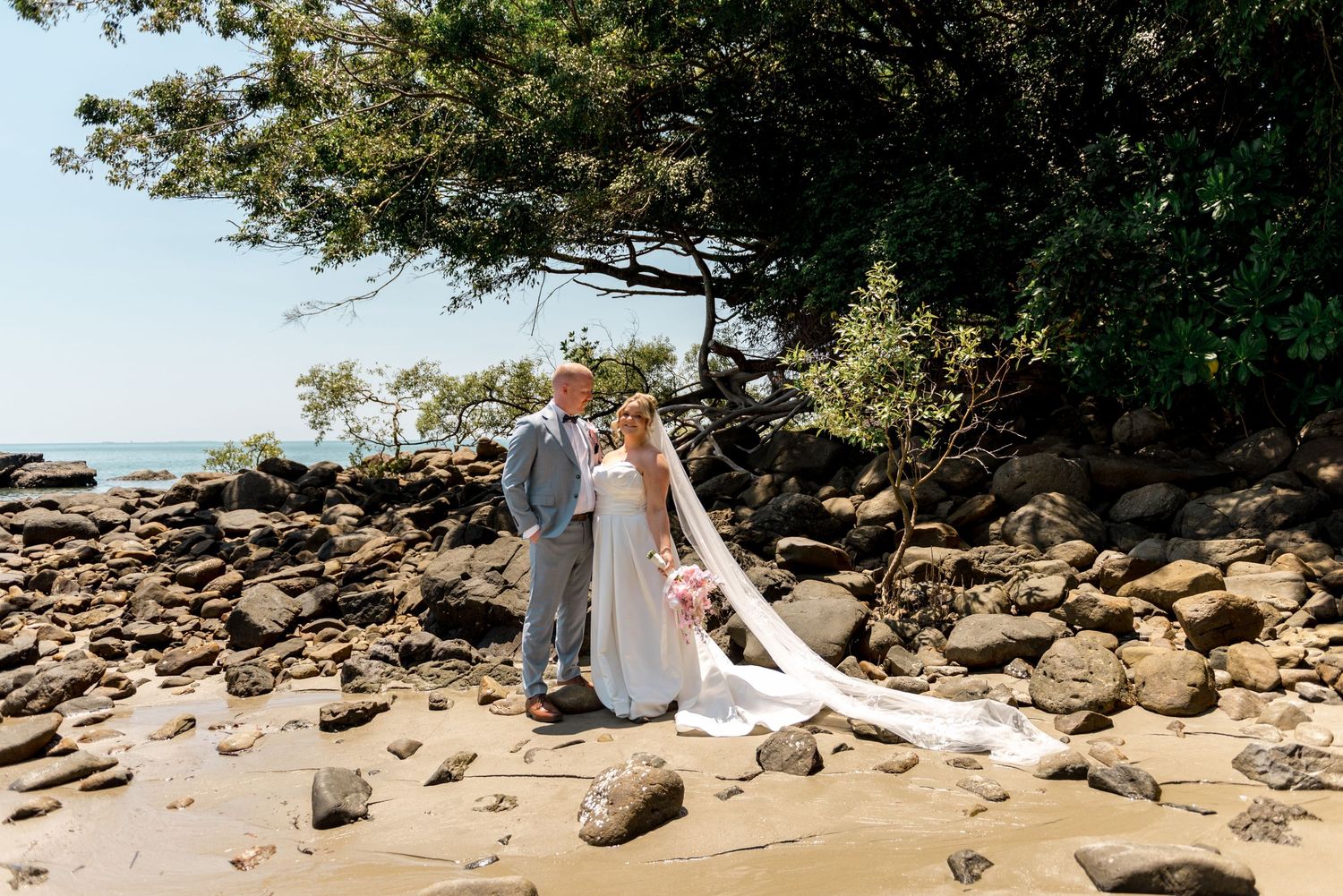Bride and groom pose together on rocky beach beside lush green trees with ocean visible behind.