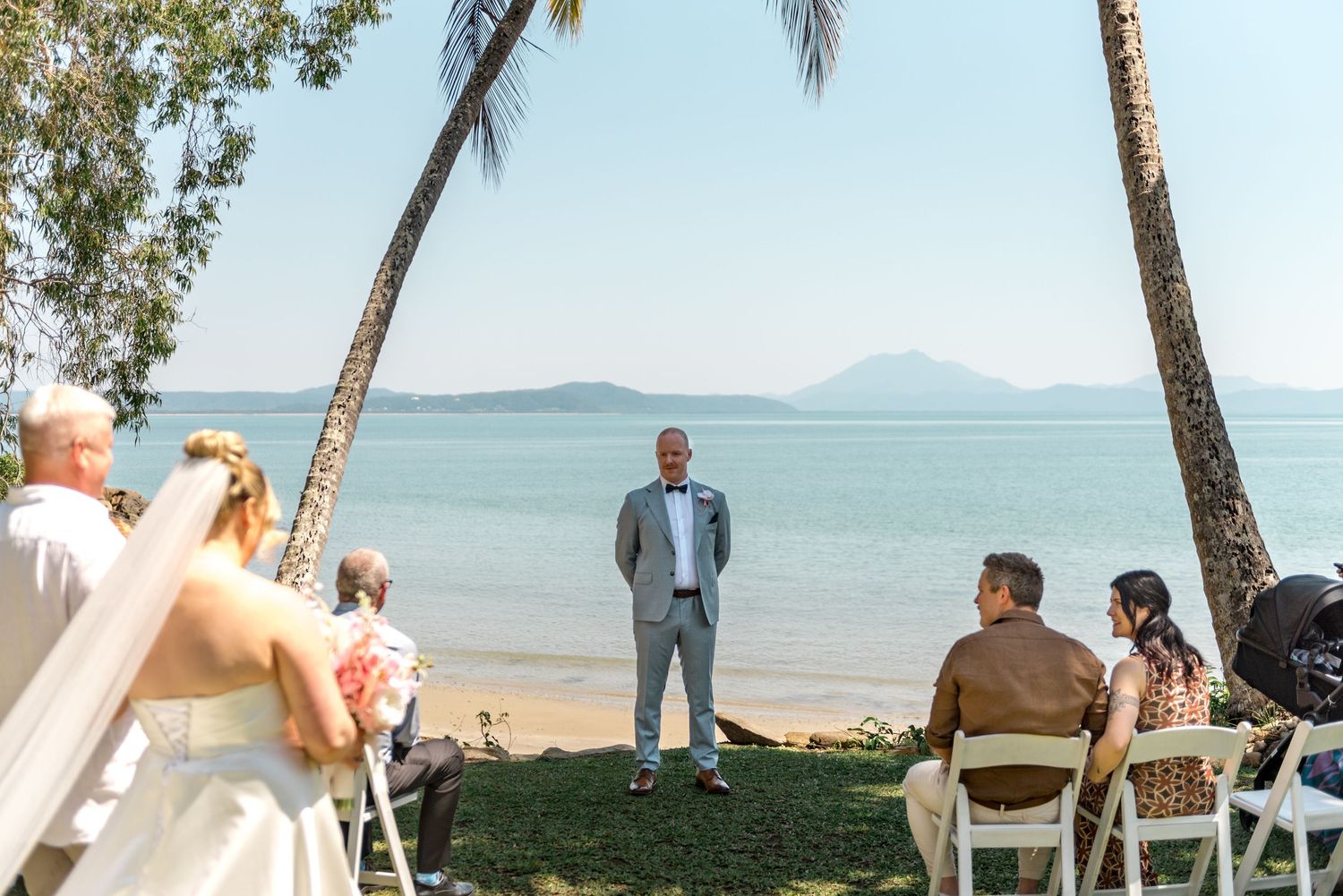 Bride walks down aisle toward groom at stunning beachfront wedding ceremony with ocean backdrop.