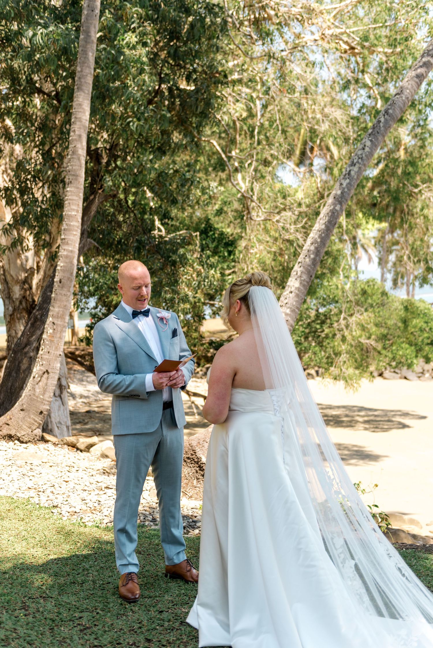 Groom holds ring box while bride looks on at tropical outdoor wedding ceremony.