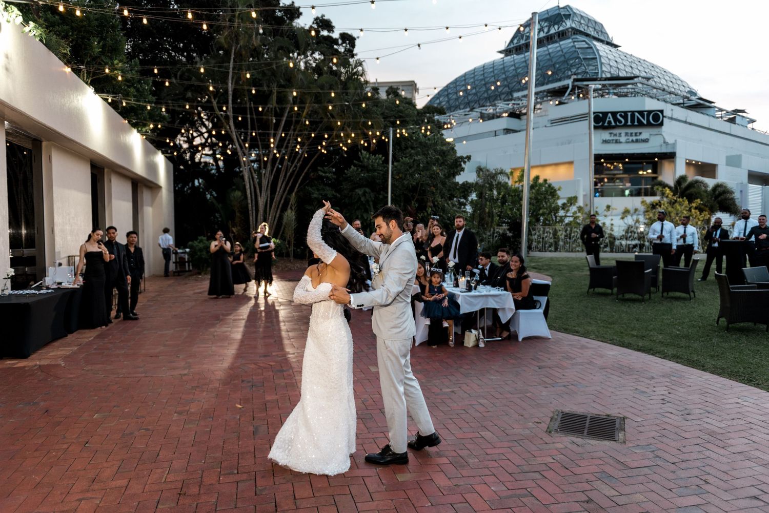 Groom twirls bride in a white wedding gown during their first dance at an outdoor evening reception with fairy lights.