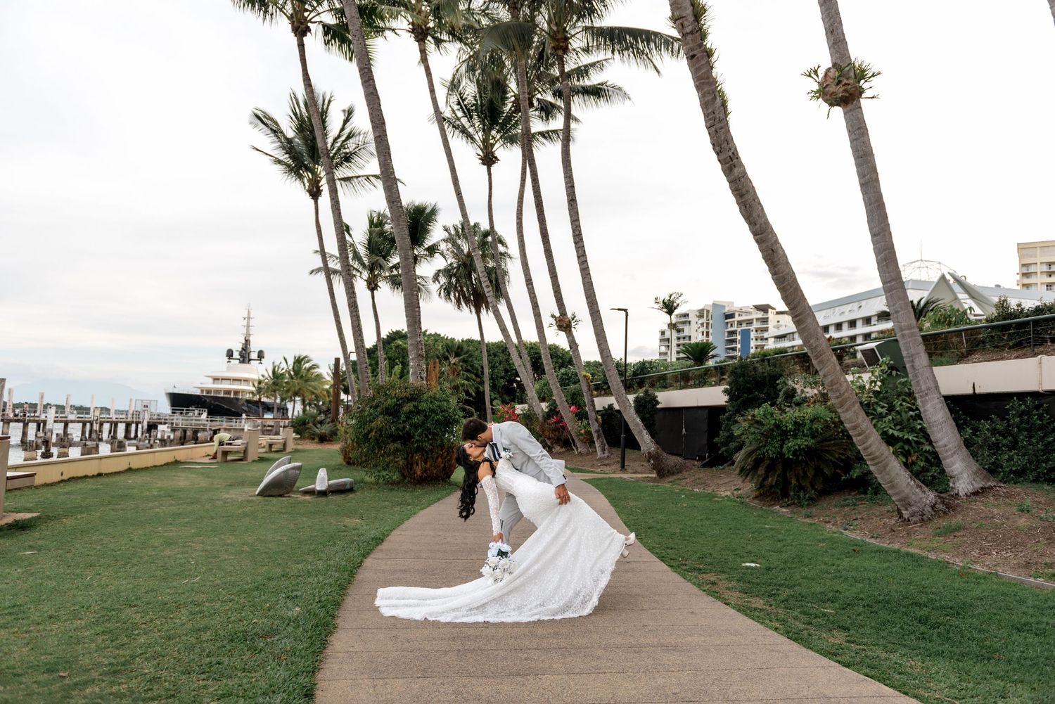Groom dips bride on curved garden path with palm trees and marina visible in background.