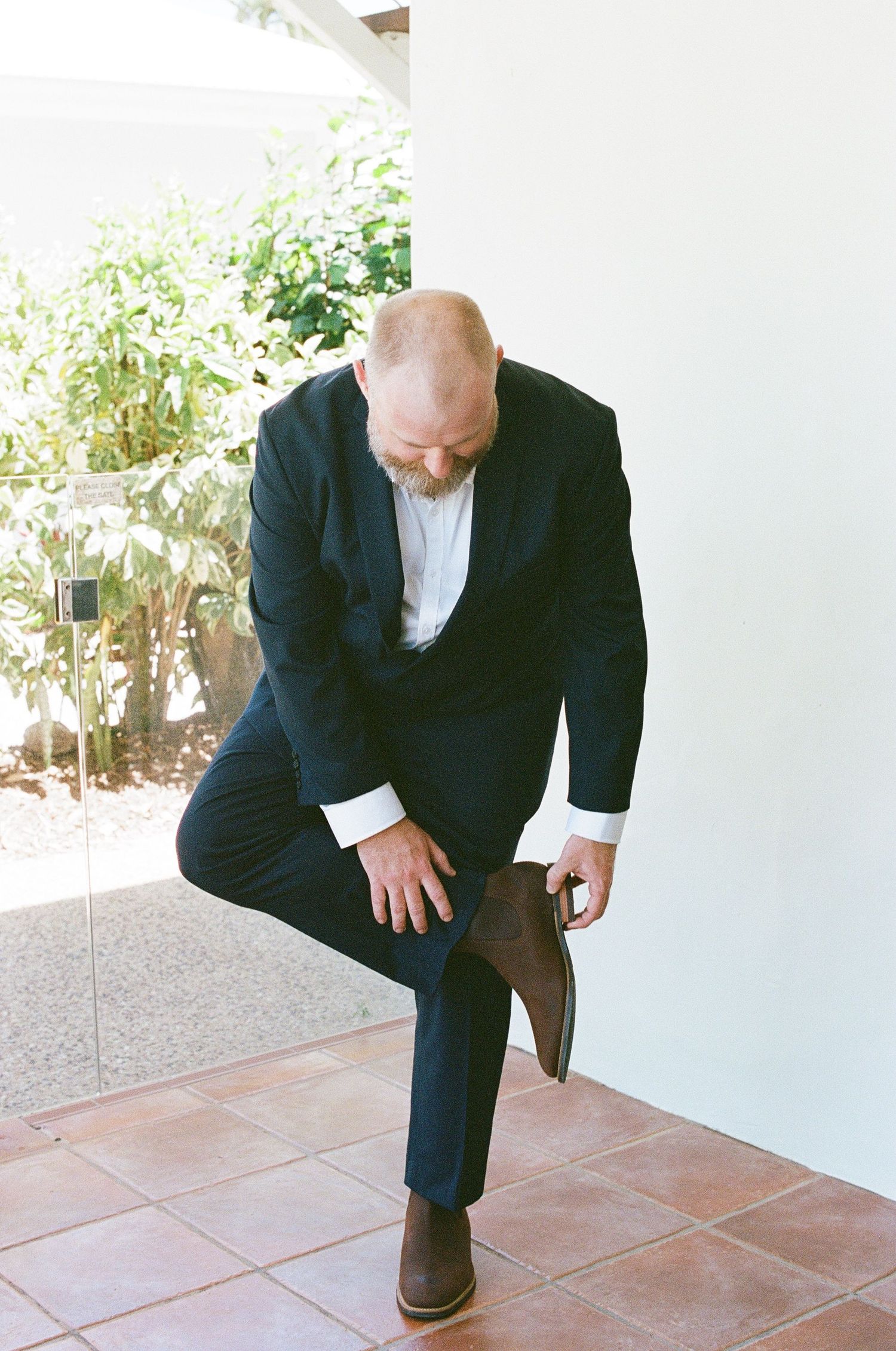 Groom in navy suit puts on brown Chelsea boots while getting ready for wedding.