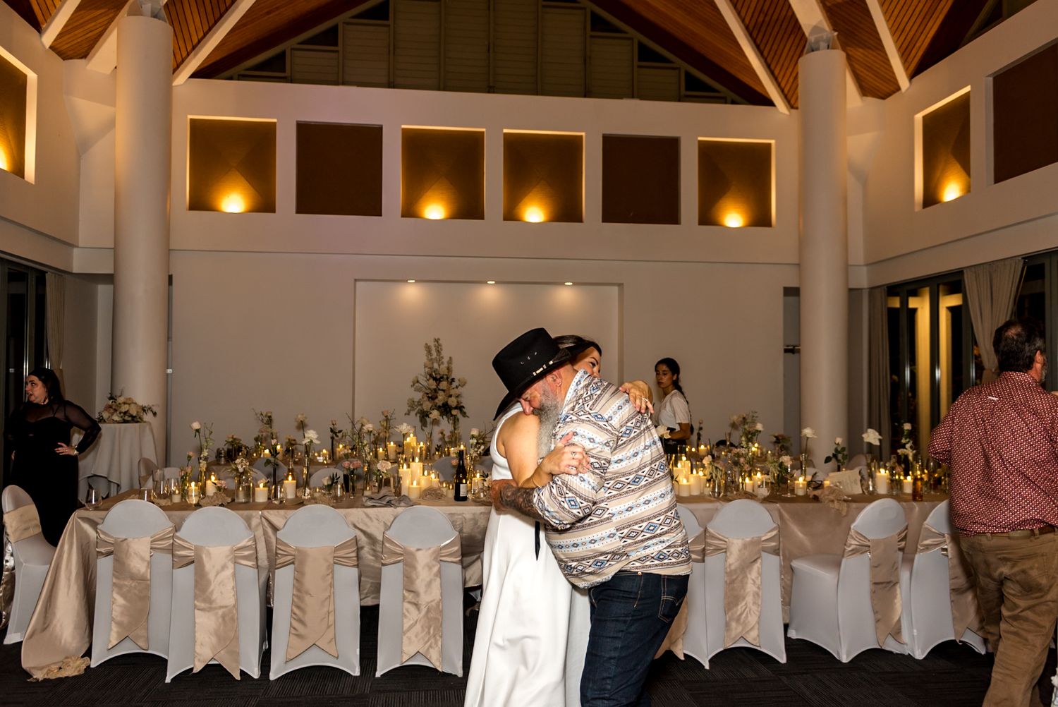 Newlyweds embracing on dance floor at wedding reception with decorated chairs and floral centerpieces.
