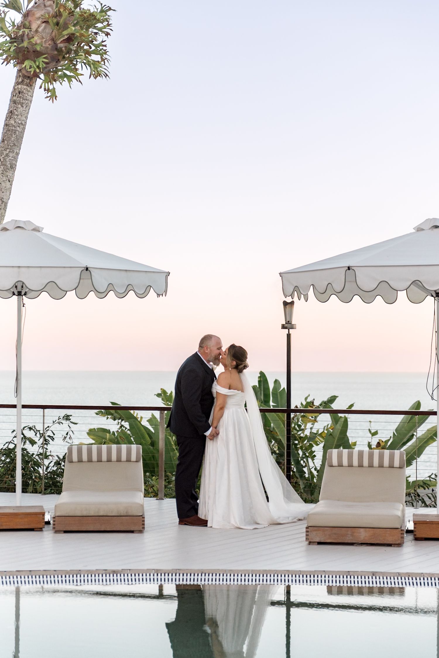 Bride and groom share a kiss by a resort pool at dusk with white umbrellas and ocean views.