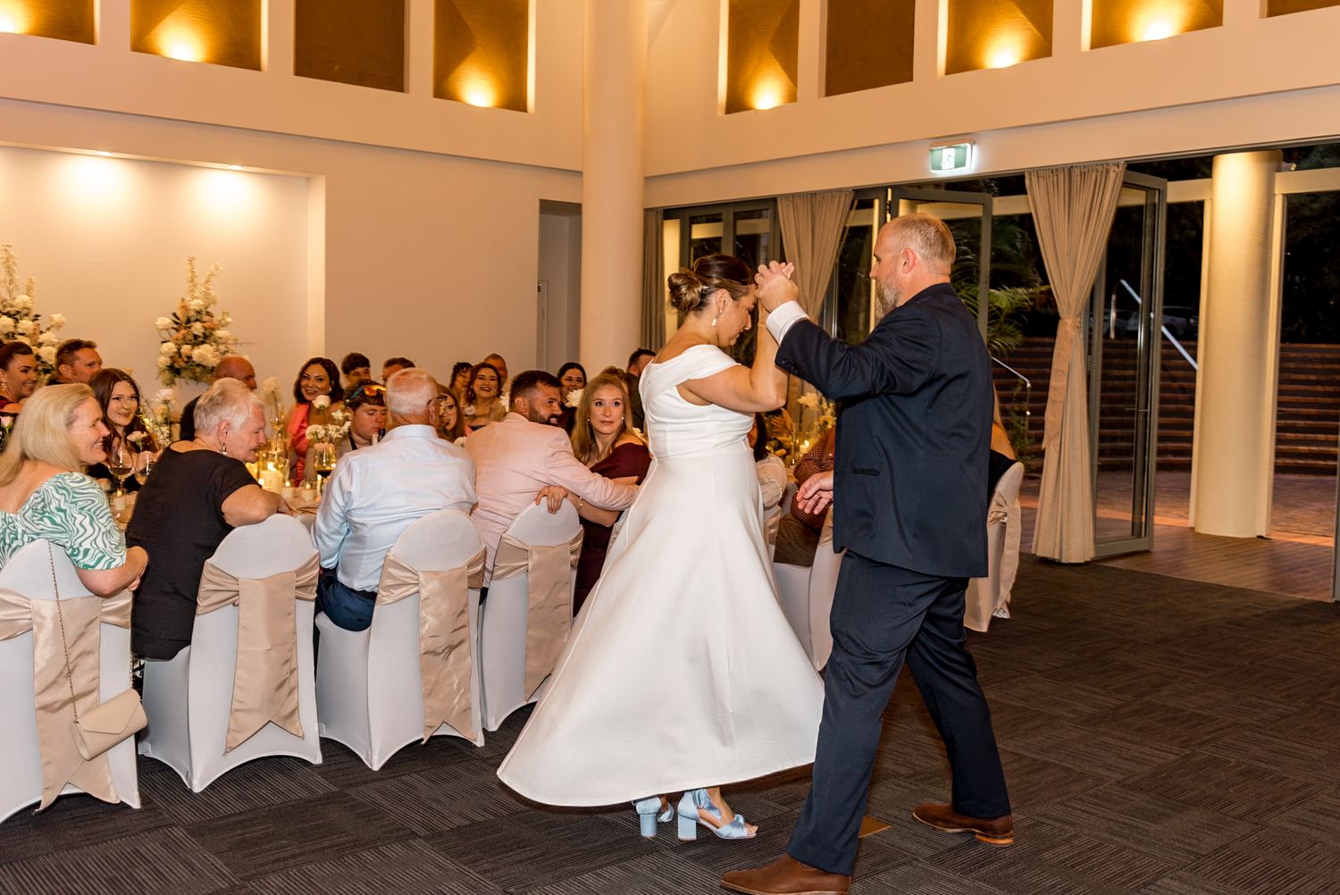 Groom twirls bride in flowing white wedding gown during first dance at elegant reception venue.