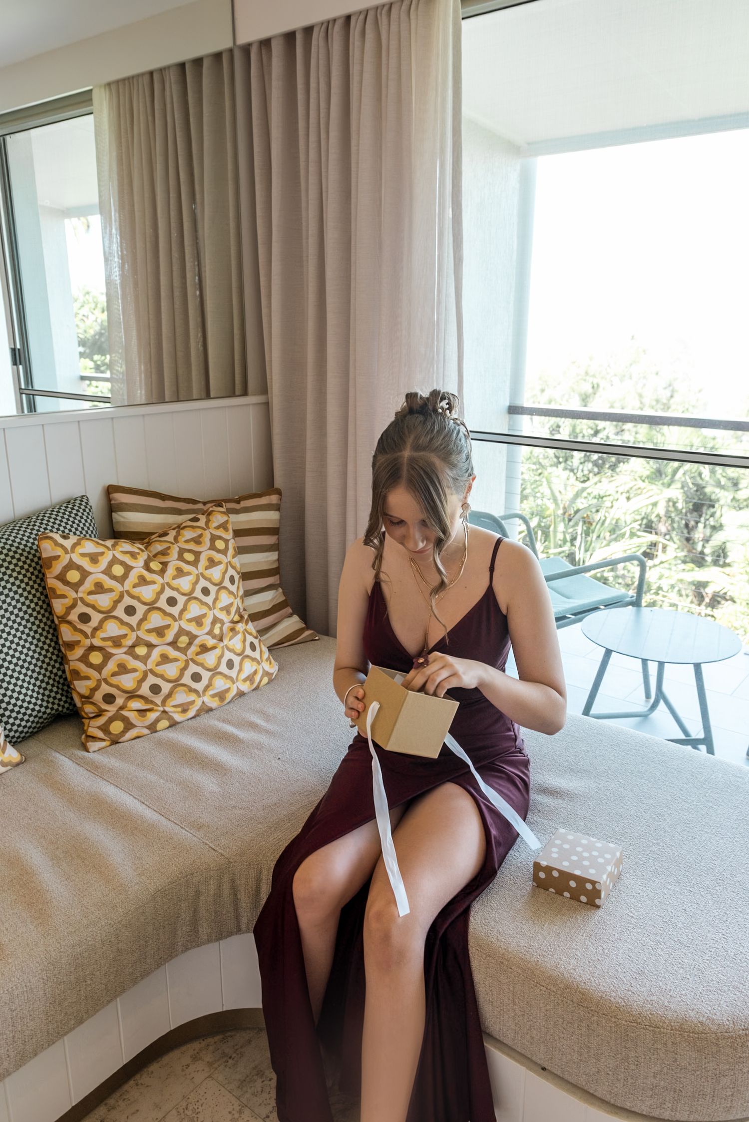 Bridesmaid in burgundy satin dress reading a card while seated on a sofa near a sunlit balcony.