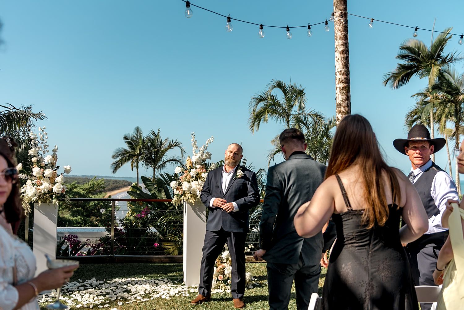 Groom awaits bride at outdoor tropical wedding ceremony with floral arch and string lights overhead.