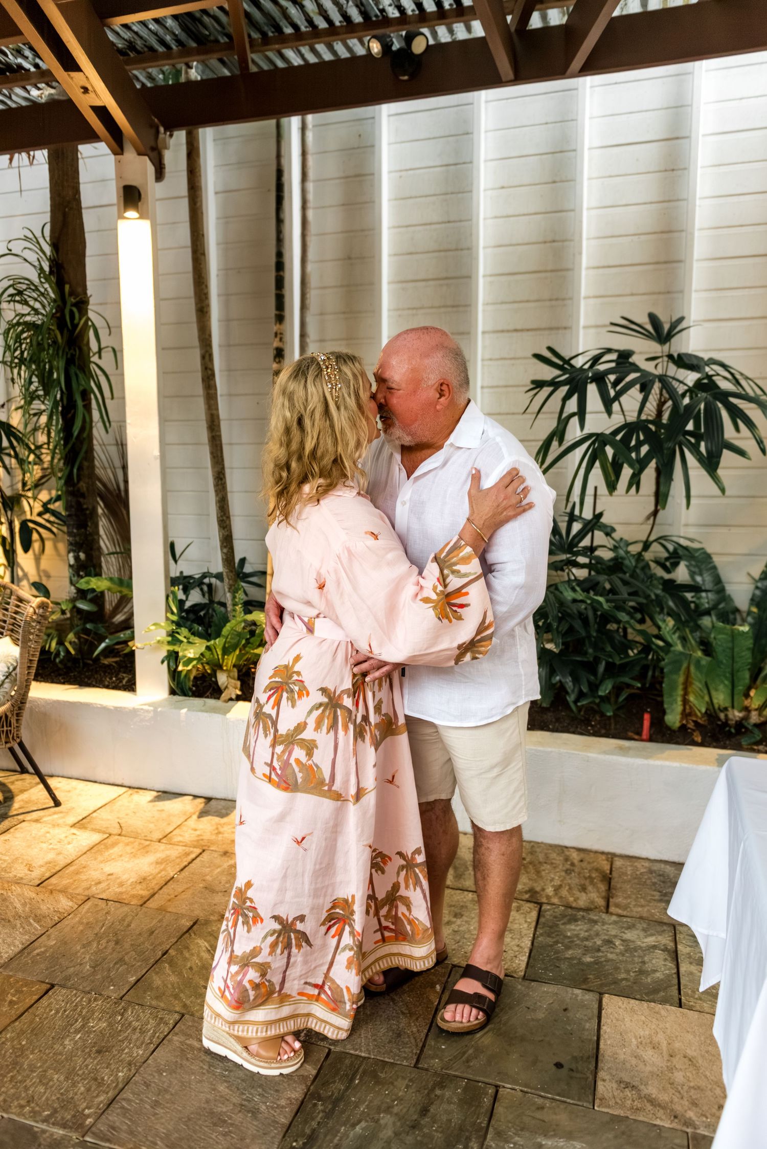 Bride and groom sharing a romantic first kiss during intimate outdoor wedding ceremony surrounded by tropical plants.
