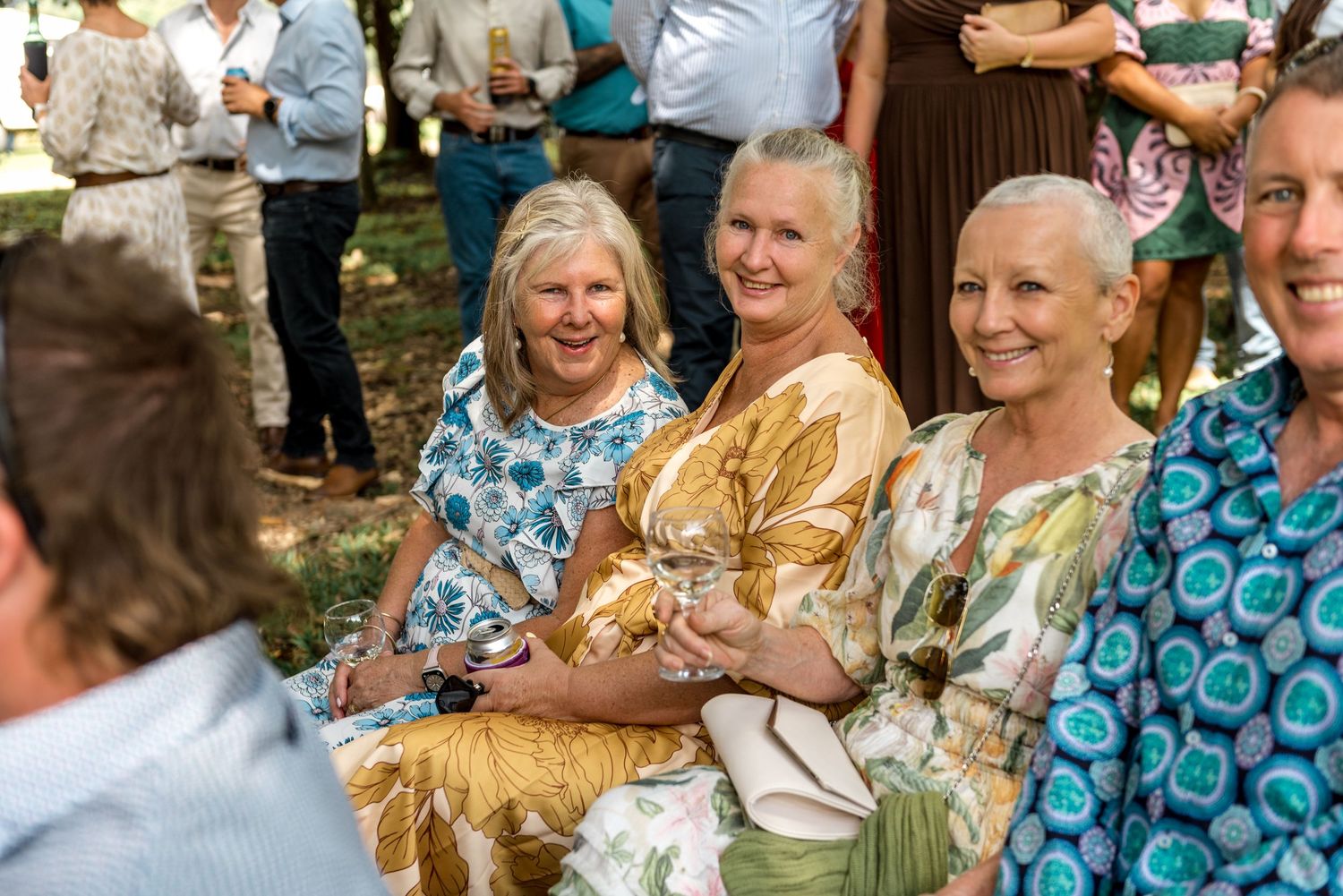 Three smiling women in floral dresses sit together as wedding guests outdoors.