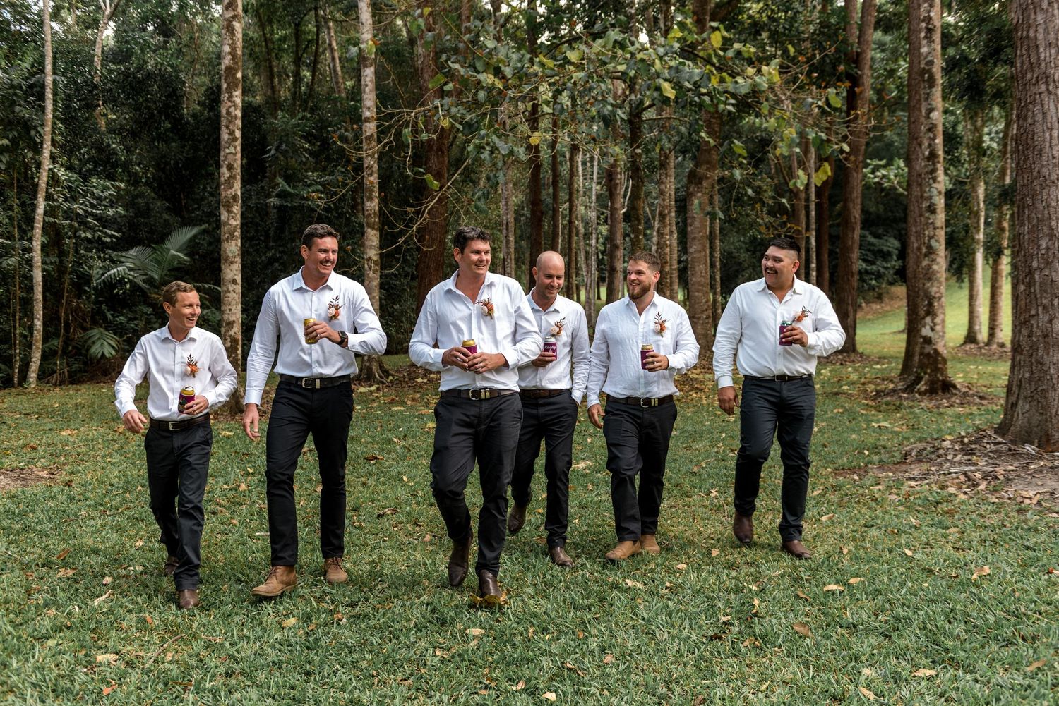 Six groomsmen in white shirts and dark pants walk together laughing through a forest wedding venue.