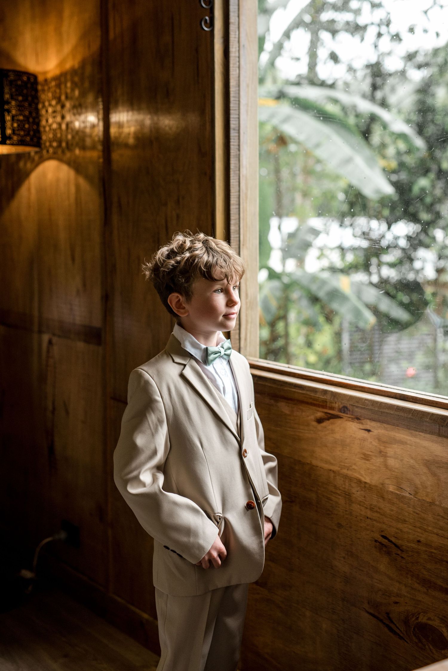 Young ring bearer in beige suit with green bow tie gazing out a wooden cabin window thoughtfully.