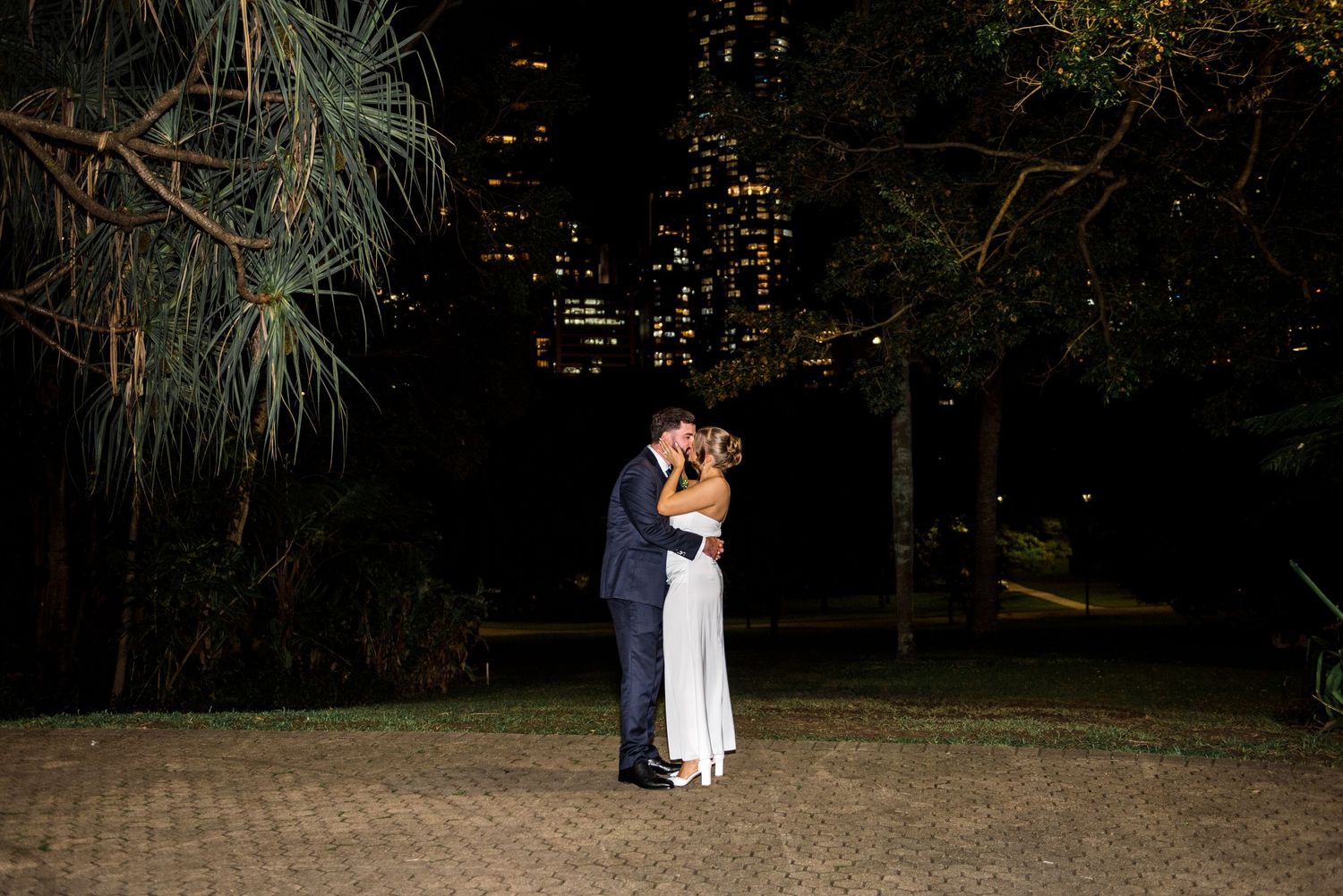 Newlyweds sharing a romantic kiss outdoors at night with city lights behind them.