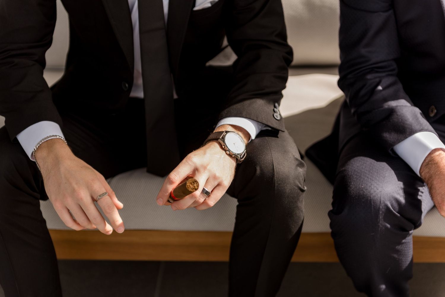 Close-up of groomsmen in black suits sitting together, one holding a cigar and wearing a brown leather watch.