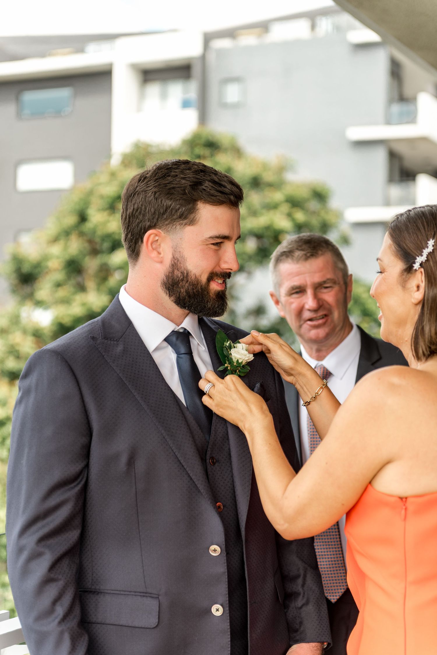 Mother in orange dress pins a boutonniere on smiling groom in navy suit while father looks on approvingly.