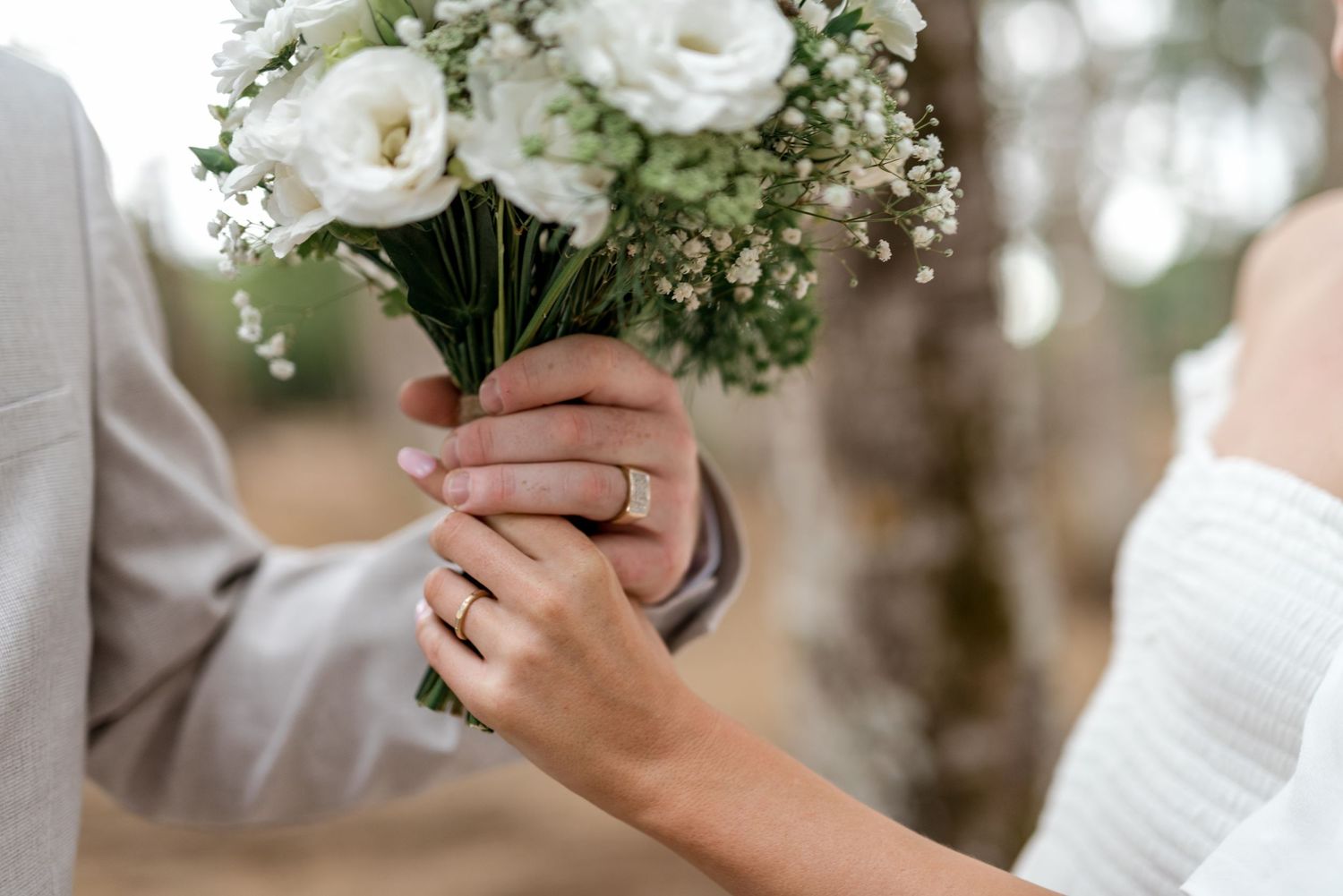 Close-up of bride and groom holding white floral bouquet with wedding rings on their hands.