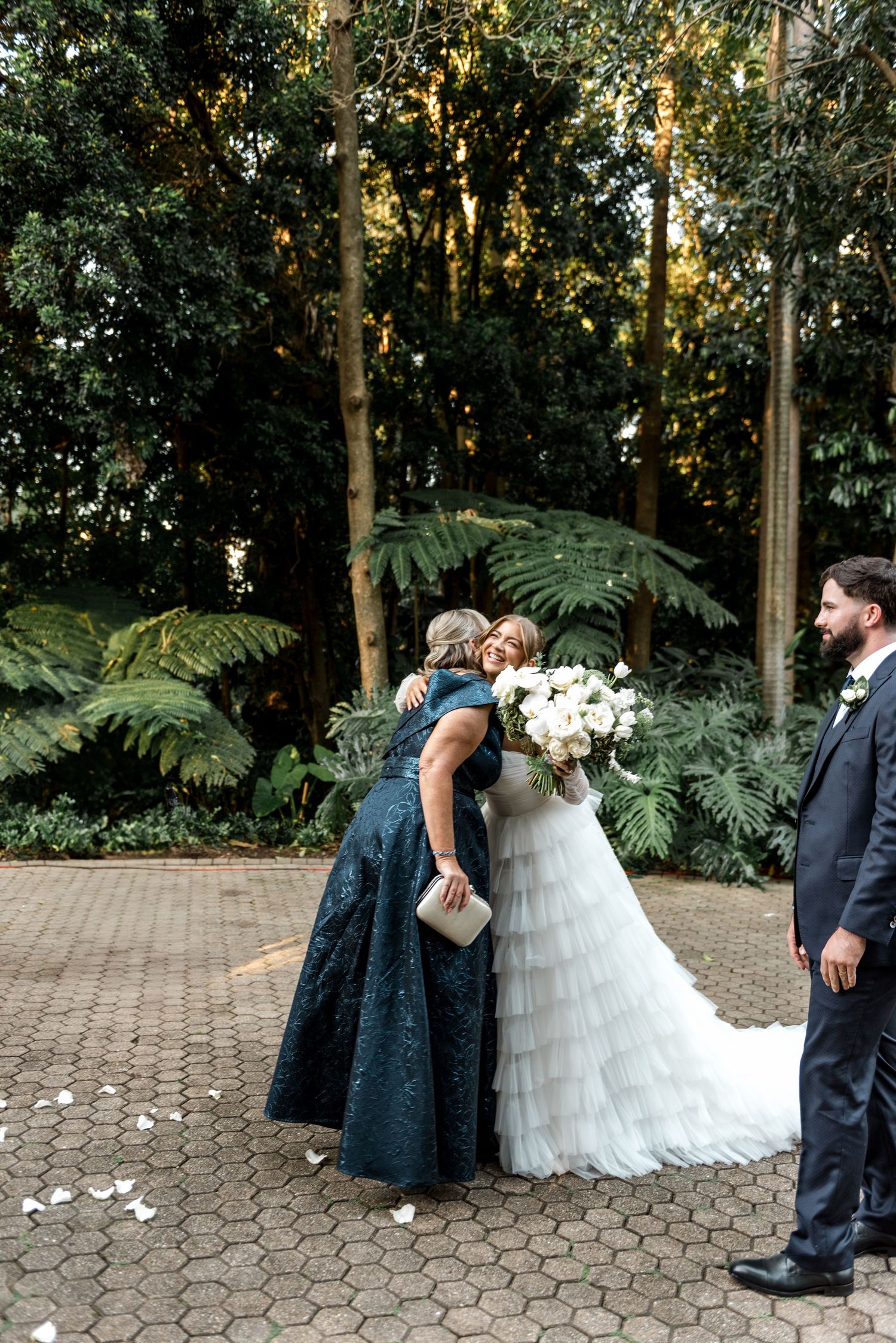 Bride embraces a woman in a teal gown while groom looks on in a lush garden setting.