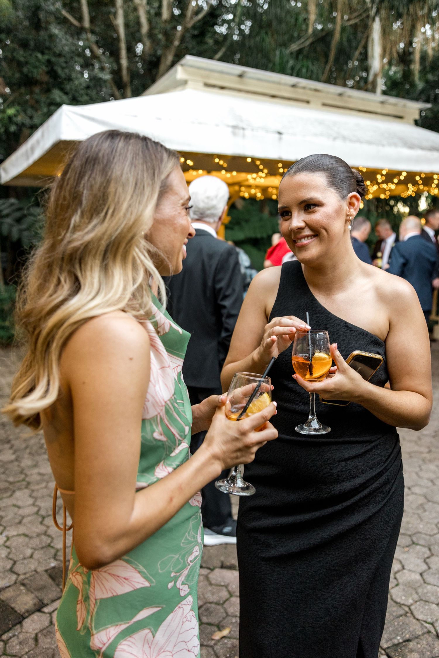 Two women in elegant attire share a cheerful conversation while holding champagne glasses at an outdoor wedding reception.