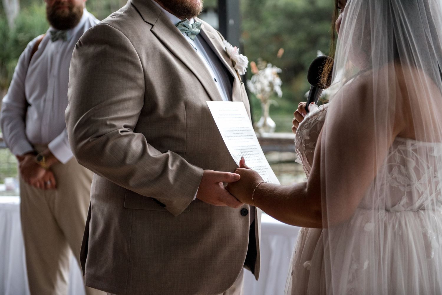 Close-up of groom in tan suit reading vows while holding brides hands during wedding ceremony.