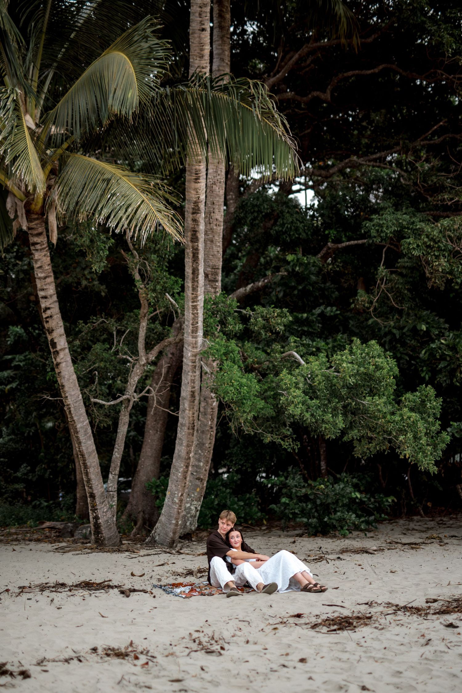 Couple relaxes together leaning against palm trees on tropical sandy beach.